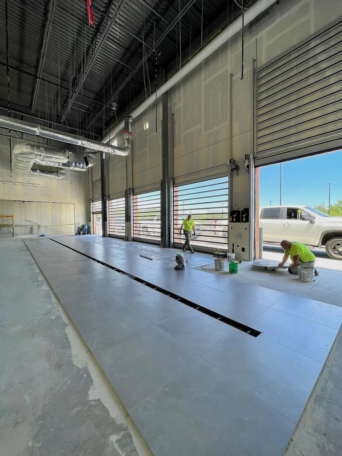 Construction workers installing large tiles on the floor of a commercial garage or warehouse with large open garage doors showing a parking lot and a white pickup truck outside.