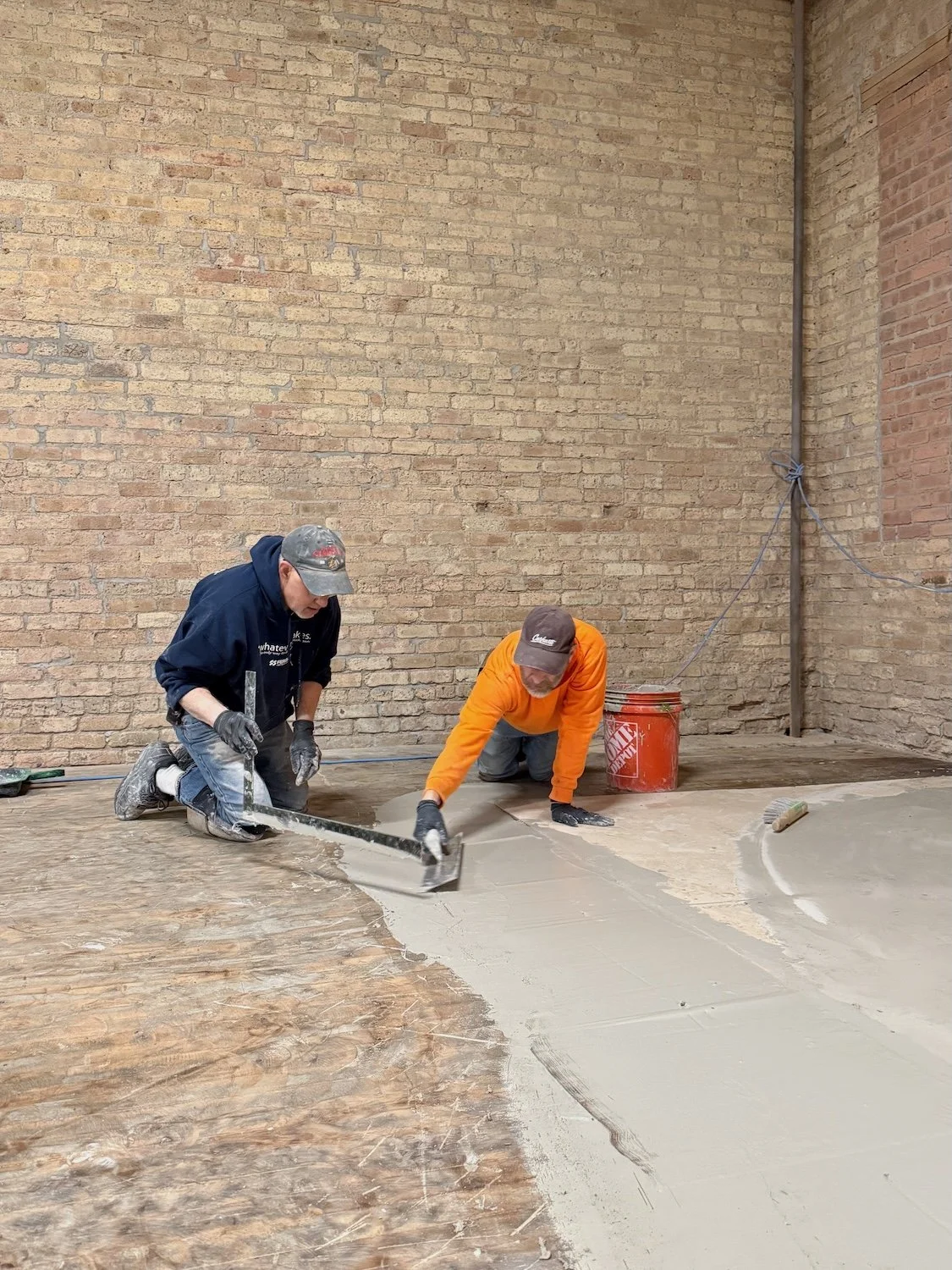 Two men are kneeling on the floor, applying concrete to a surface inside a room with brick walls. One man is spreading the concrete with a trowel, while the other is smoothing it with a hand tool.