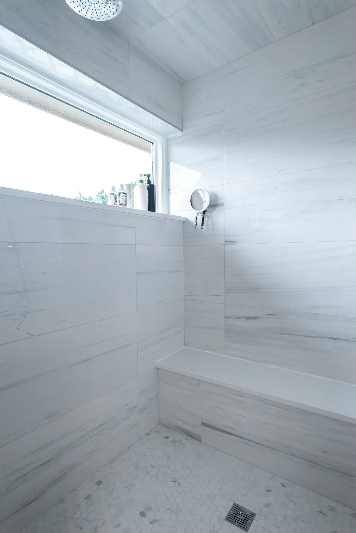 A modern, minimalistic shower with white marble tiles on the walls and small hexagon tiles on the floor. A window with natural light, toiletries on the ledge, and a round showerhead.