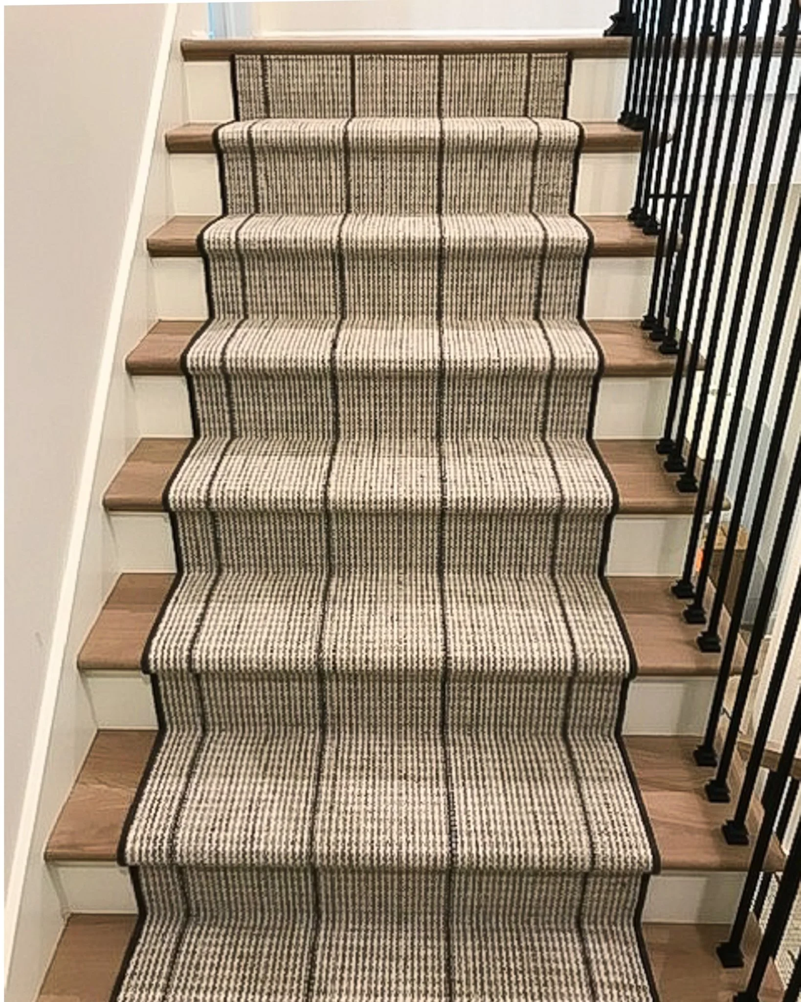 Staircase with a striped beige and black carpet, wooden steps, white walls, and a black metal railing on the right side.