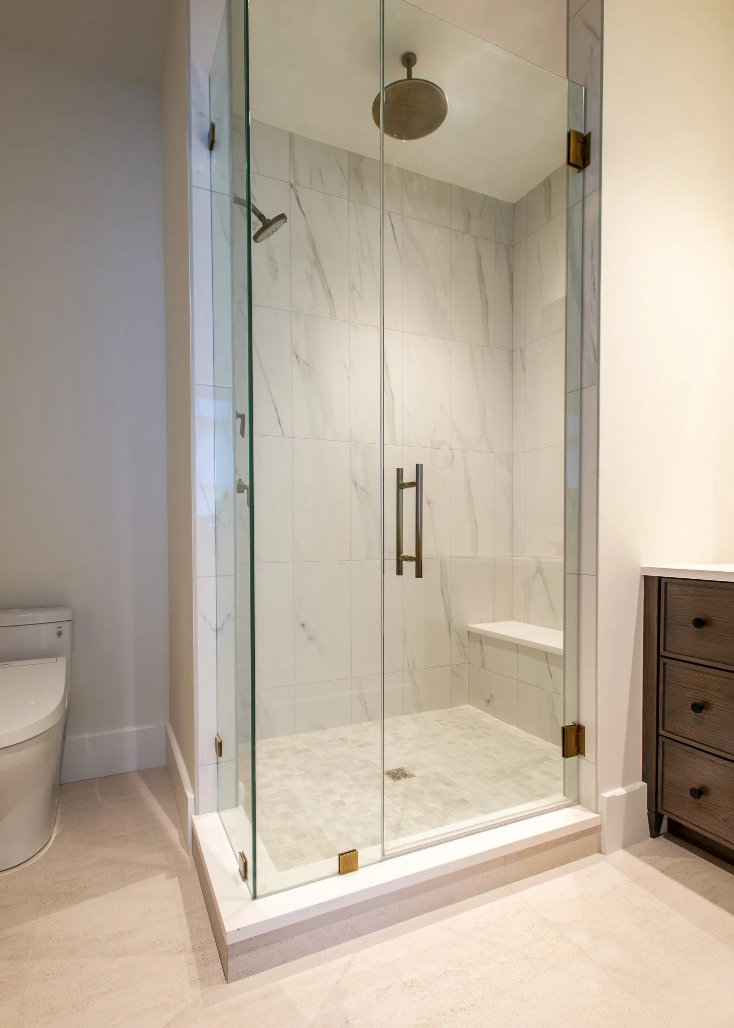 A walk-in shower with glass doors, white marble tiles, a rainfall showerhead, and a handheld shower. There is a small bench built into the wall and a metal handle on the door. To the right, there is a wooden cabinet with drawers. The surrounding bath