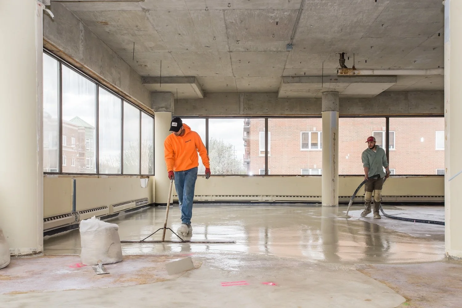 Two construction workers are applying a concrete floor coating inside an unfinished building. One worker is wearing an orange hoodie and gloves, using a long-handled tool. The other worker, in a grey shirt and red cap, is handling a hose. Large windo