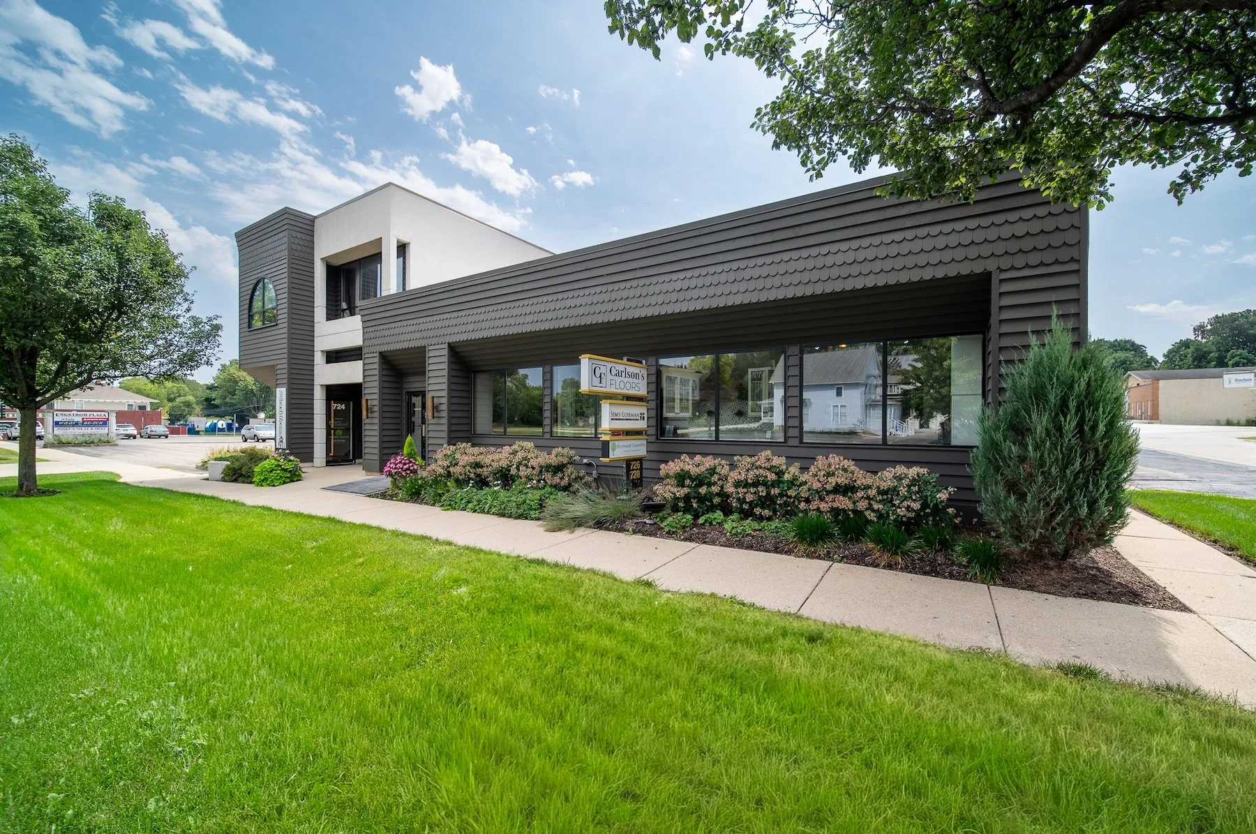 A modern commercial building with dark siding, large front windows, and landscaping with bushes and flowers in front. The building has a sign for Carlsons Floors and is situated on a well-maintained lawn with a sidewalk and parking lot nearby, under a partly cloudy sky.