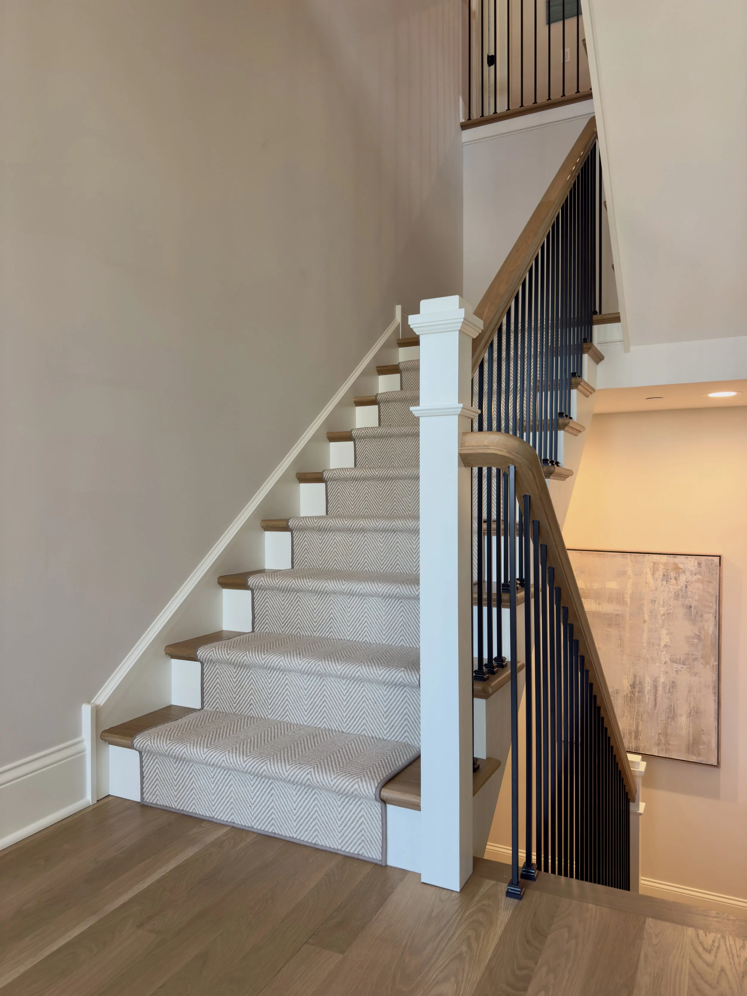 Interior view of staircase with carpeted steps, wooden handrail, and black metal balusters, adjacent to painted walls and hardwood flooring.