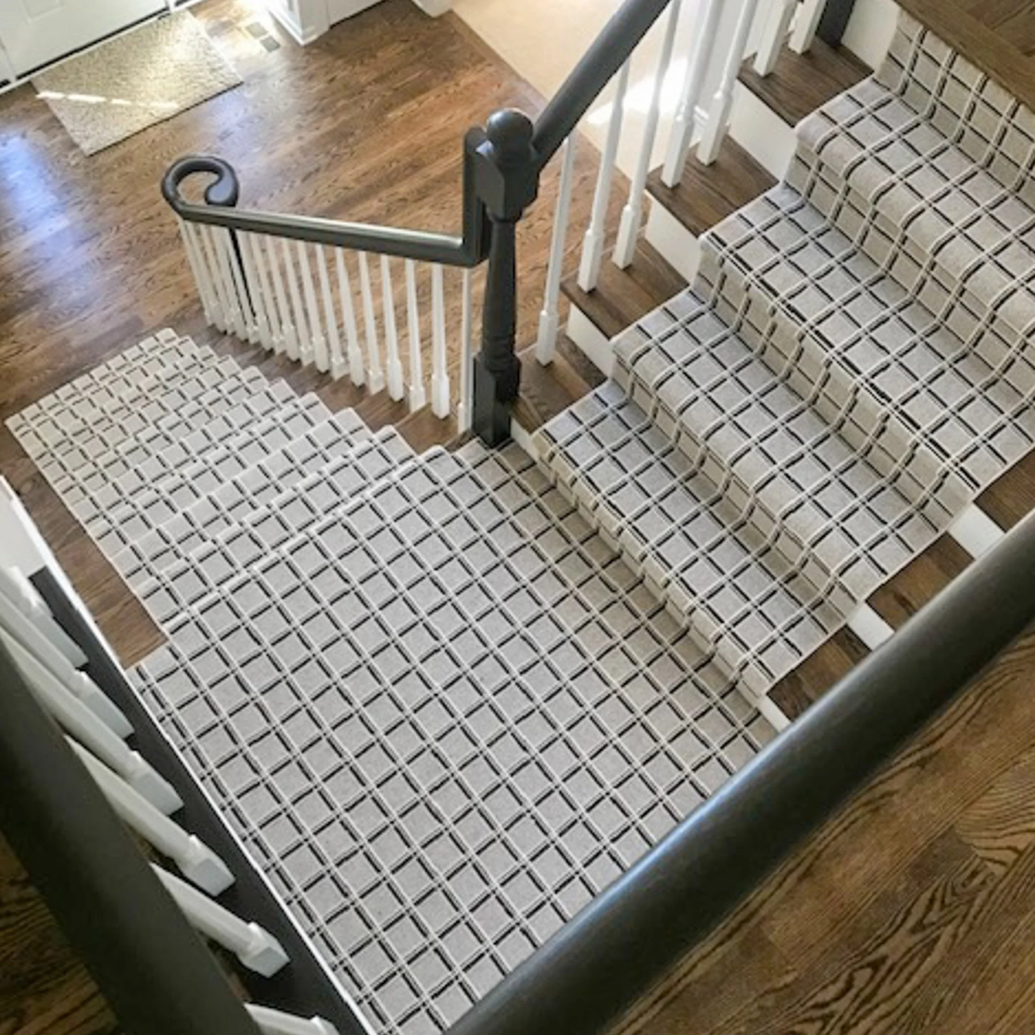 View of a staircase with a black handrail, white balusters, and a checkered carpeted stair tread. A wooden floor is visible at the top of the stairs and a beige mat on the floor near a door.