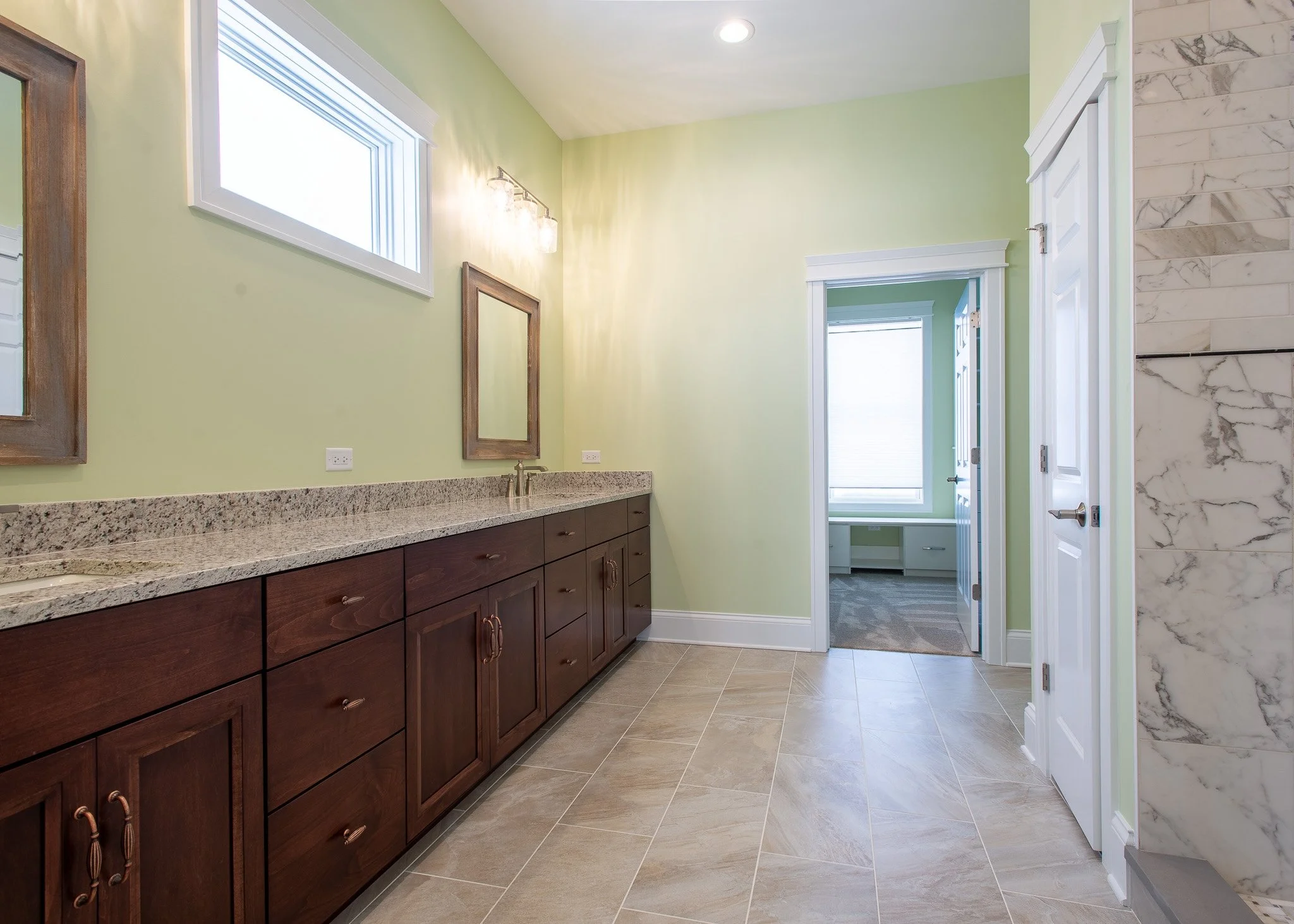 A bathroom with a green wall, brown wooden cabinet with drawers, granite countertop, marble tiled wall on the right, and a doorway leading to another room with a window.