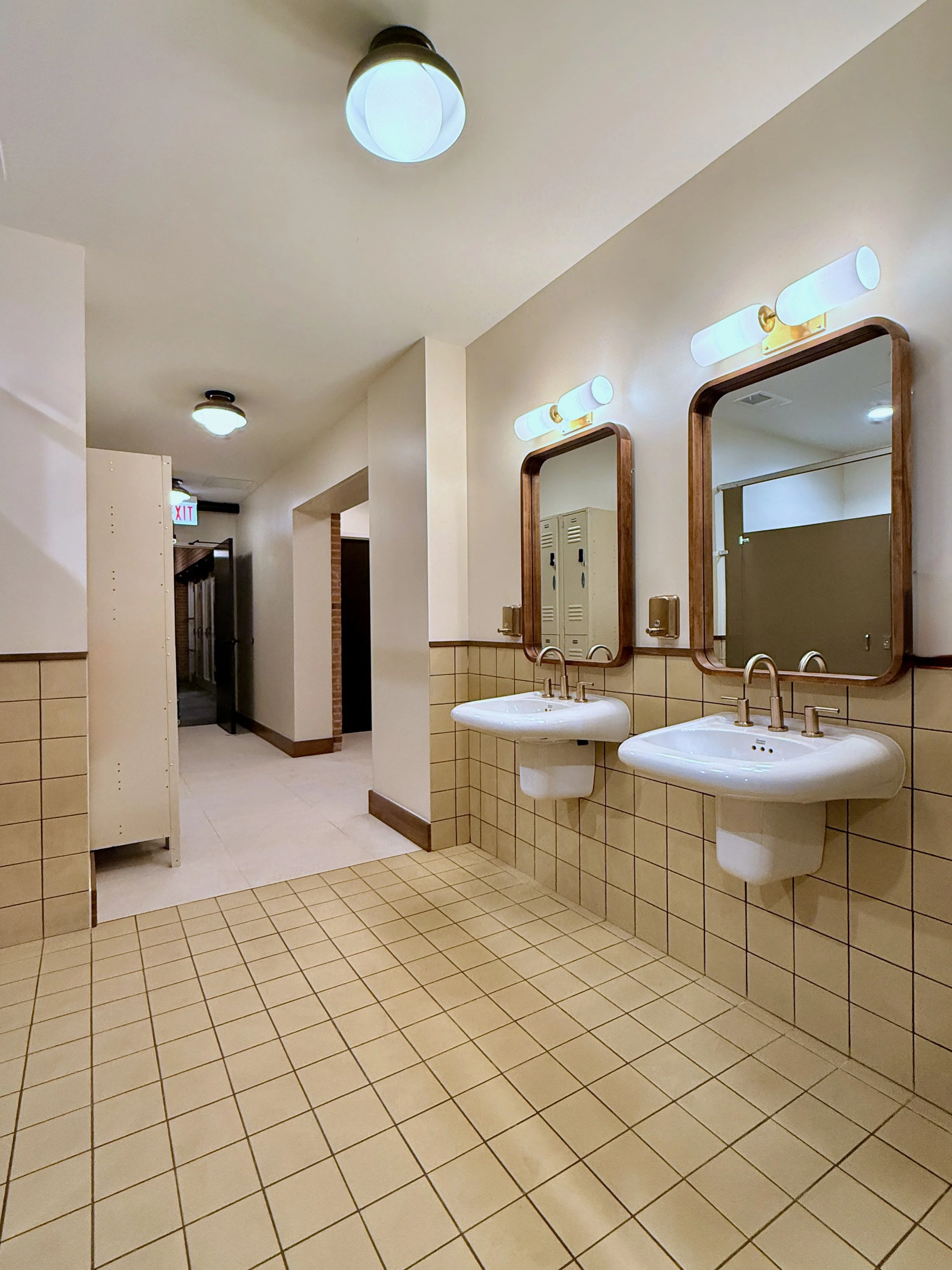 Restroom with two sinks and mirrors, beige tile walls and floor, ceiling lights, lockers, and an exit sign.