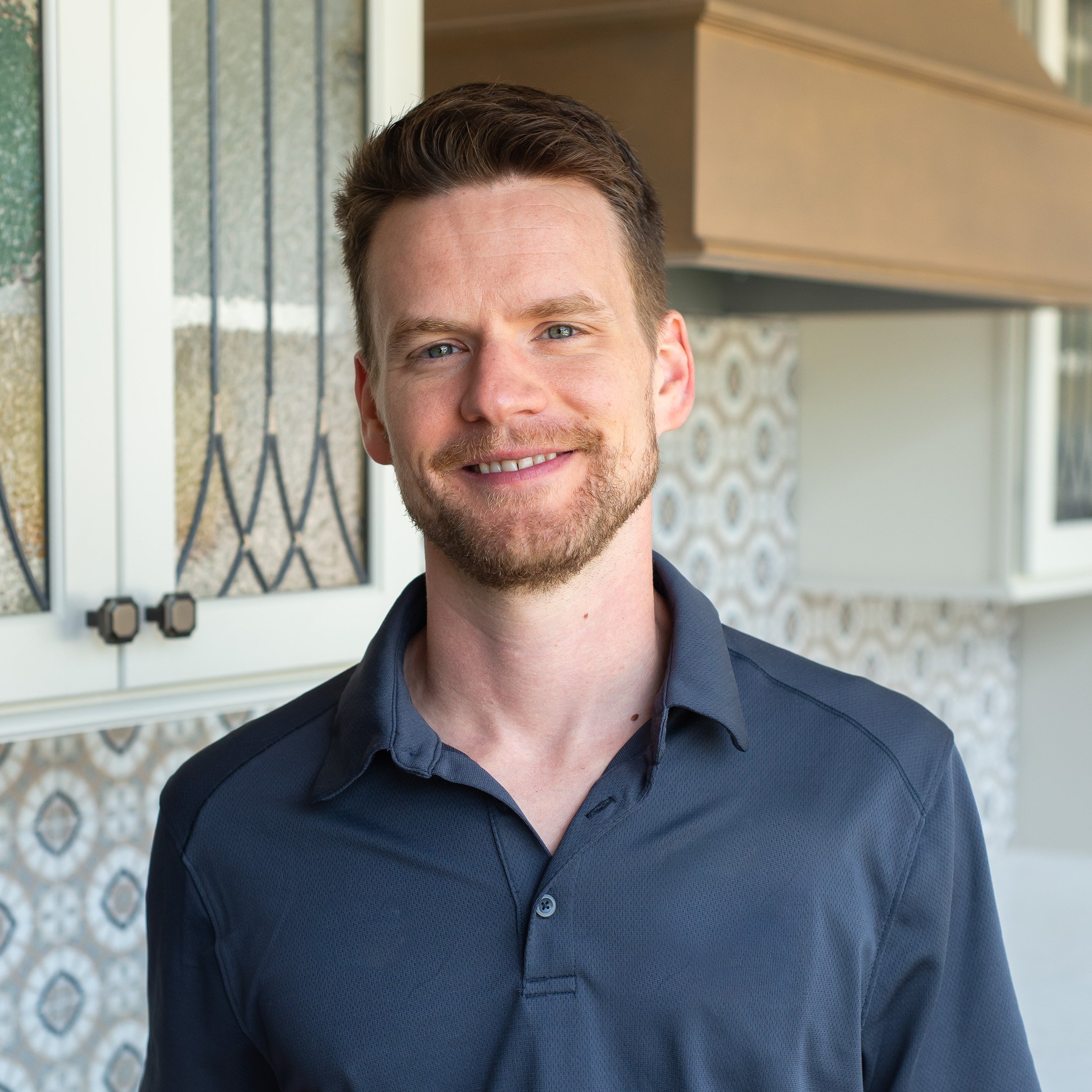 Close-up of a young man with light skin, short brown hair, and blue eyes, smiling, wearing a dark blue button-up shirt, standing outside in front of a beige house with patterned siding.