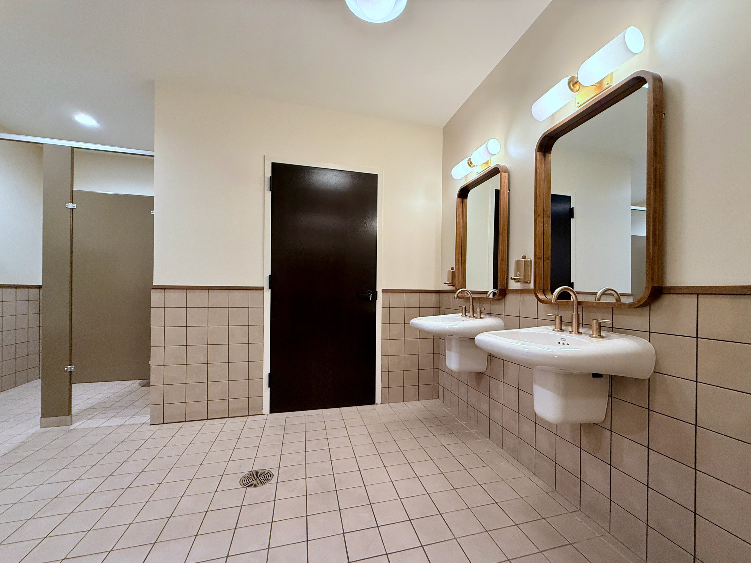 A restroom with three sinks and three mirrors above them, beige walls, and tiled floors. There are two light fixtures above the mirrors, and a door with a dark finish on the wall.
