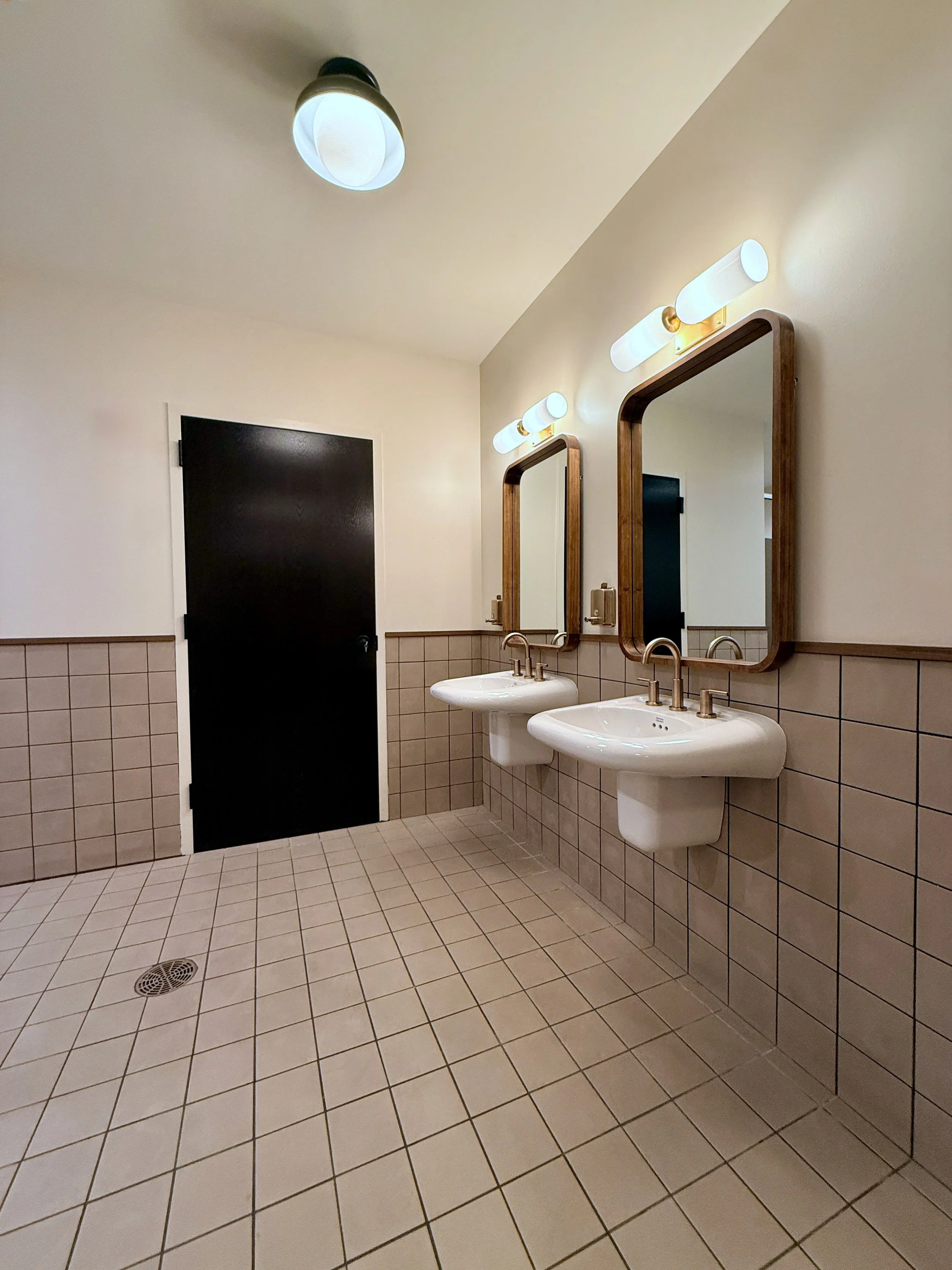 Public restroom with two sinks, wooden framed mirrors, white tiled walls, and beige tiled floor.