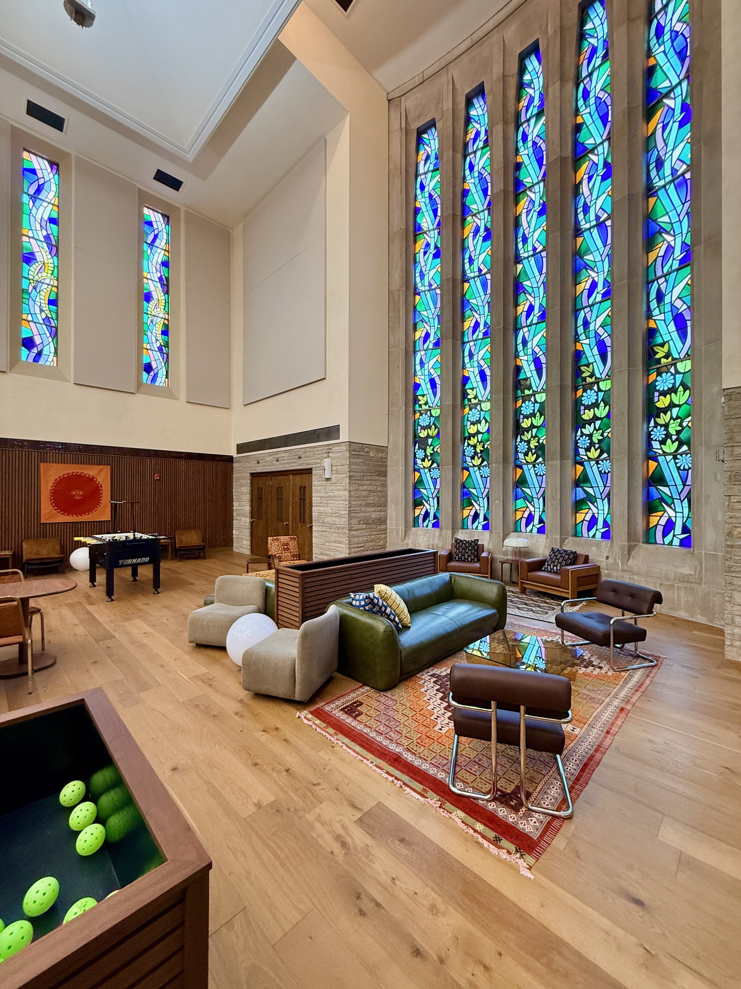 Interior of a spacious room featuring stained glass windows, modern chairs, a green sofa, a patterned rug, and a table with green balls, suggesting a recreational or lounge area.