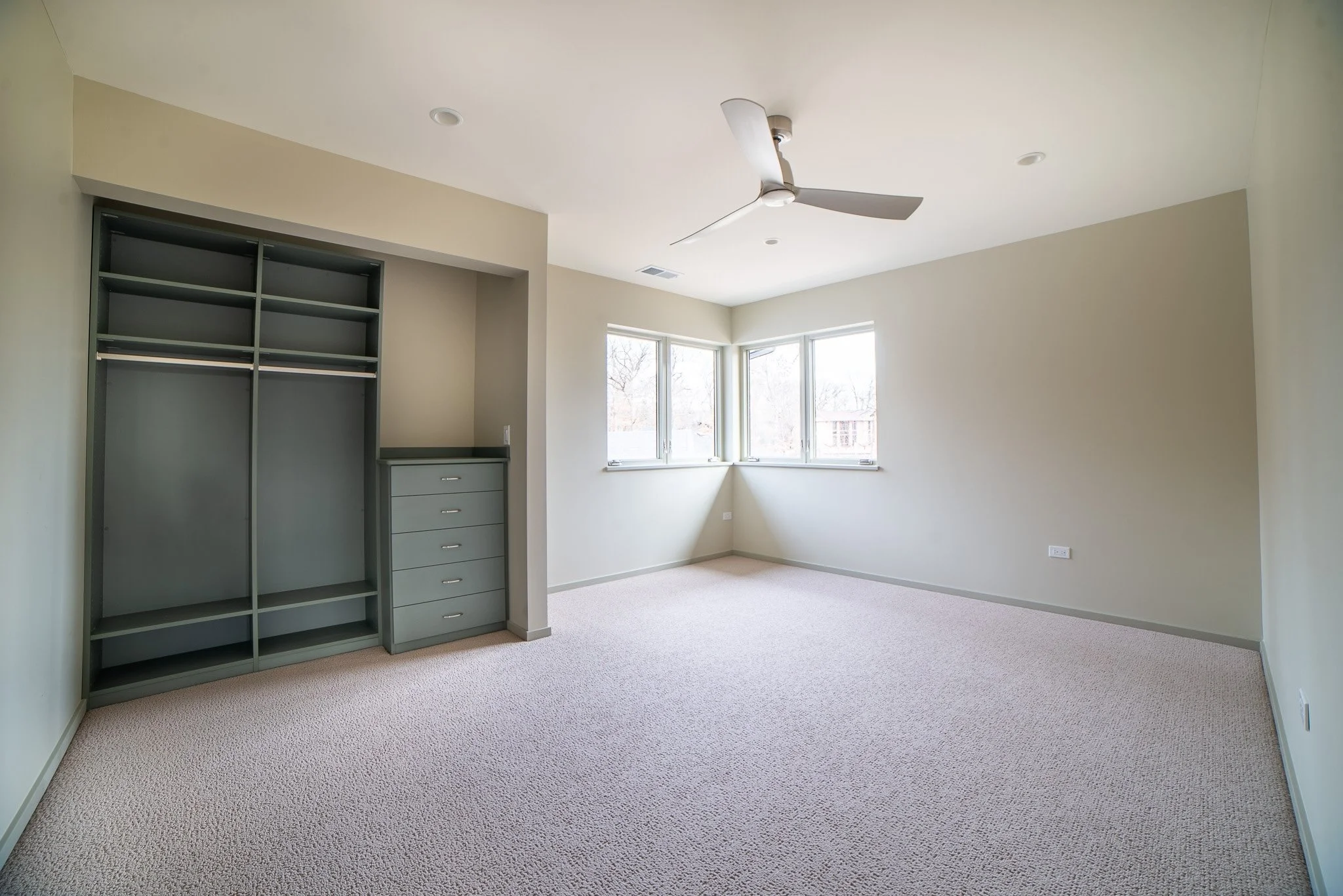 Empty bedroom with beige carpet, cream walls, a window with three panes, a ceiling fan, and an open closet with shelves and drawers.