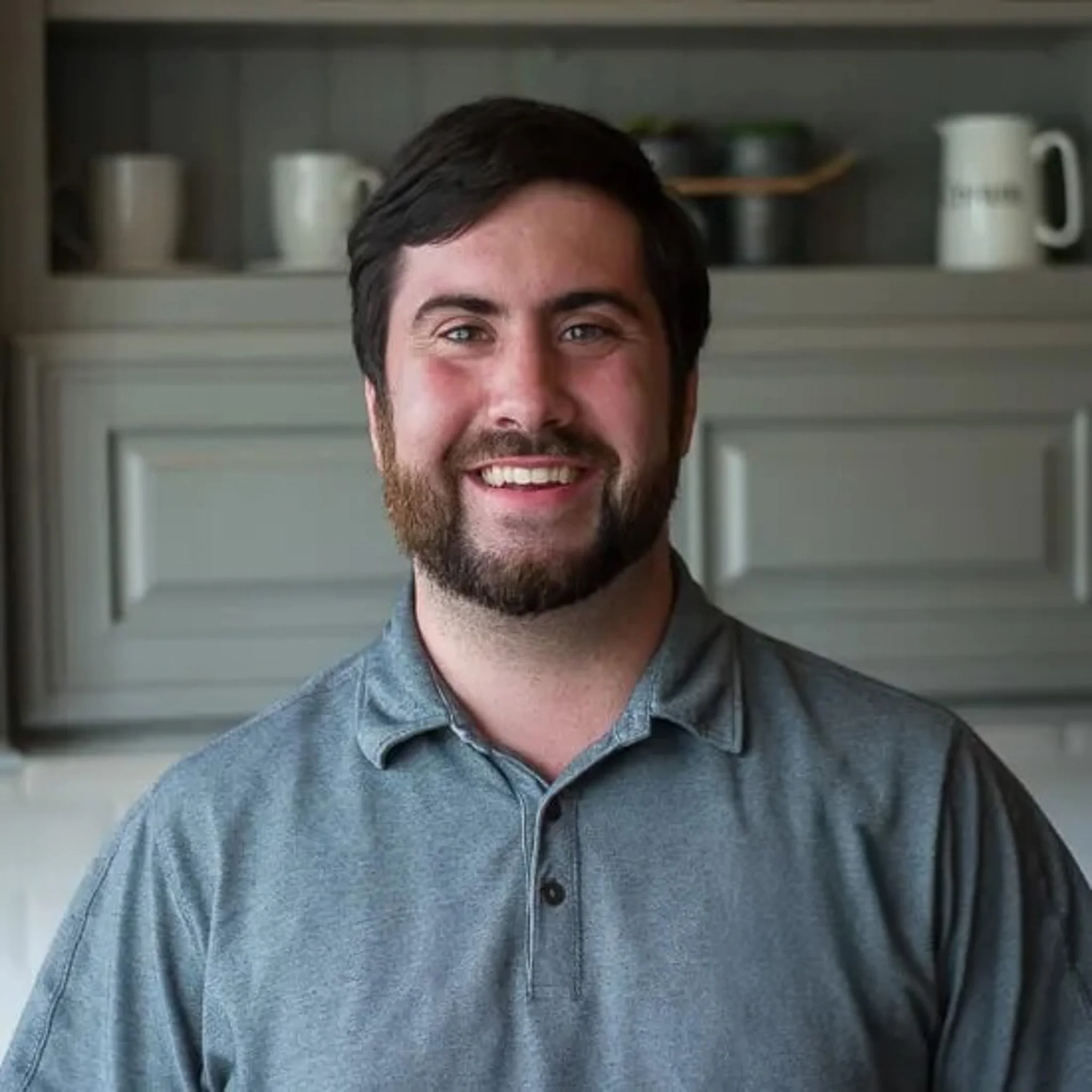 Close-up of a smiling man with dark hair and a beard, wearing a grey collared shirt, standing in front of a cabinet with cups, pitchers, and kitchen items.