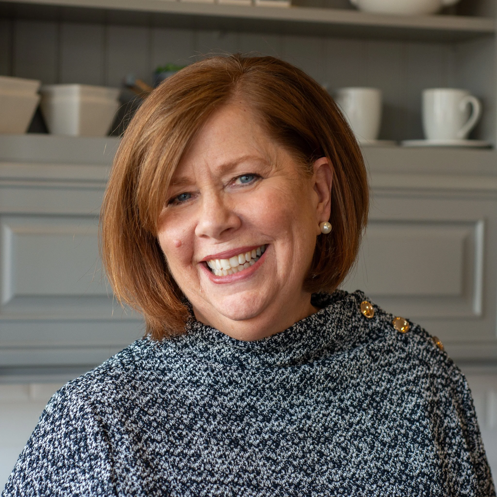 Close-up of a woman with red hair, blue eyes, and wearing pearl earrings, smiling in an indoor setting with shelves of white cups and containers in the background.