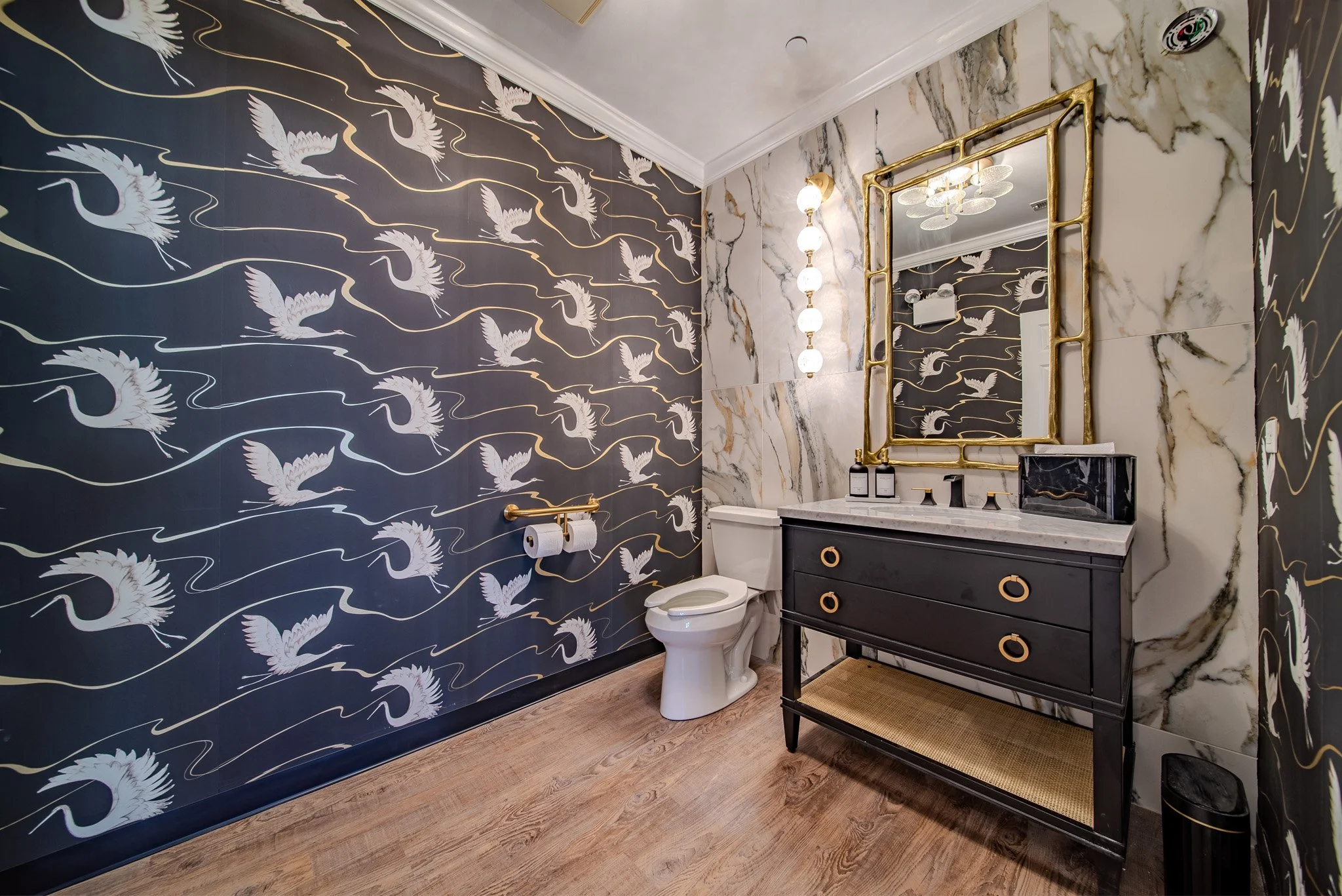 A luxurious bathroom featuring dark wallpaper with white cranes and gold accents, a marble wall behind the mirror, a white toilet, a black vanity with gold handles, and a gold-framed mirror.