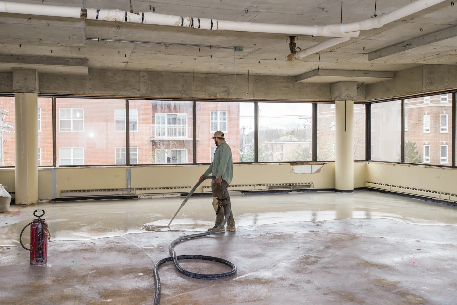 A construction worker in a hard hat and green jacket is working on a concrete floor inside a large room with floor-to-ceiling windows. He is holding a hose, pouring or spraying a liquid on the floor. The room is under renovation, with exposed pipes o