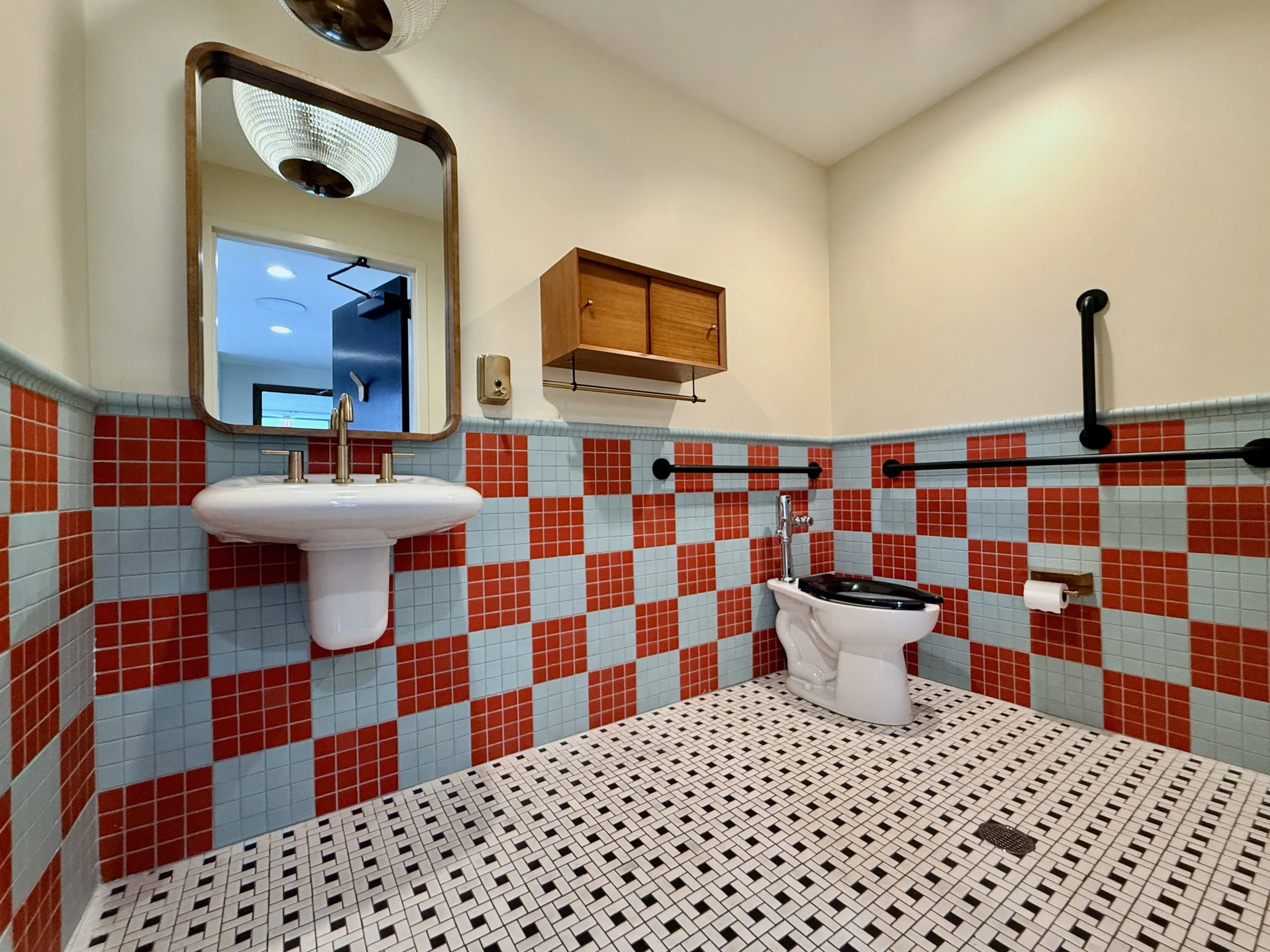 Bathroom with orange, light blue, and white tiled walls, a mirror, a white pedestal sink, a wall-mounted wooden cabinet, black grab bars, a toilet, a toilet paper holder, and a tiled floor with a drain.