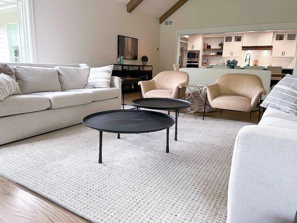 Living room with a large beige sofa, two beige armchairs, two black circular coffee tables, a flat-screen TV, and a white area rug.