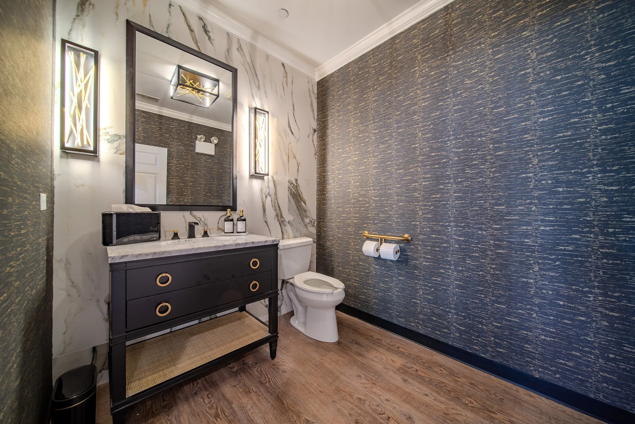 A modern bathroom featuring a sleek black vanity with gold ring handles, a white marble countertop, a large rectangular mirror, gold and black fixtures, a toilet with a dual toilet paper holder, dark textured wallpaper, and wood flooring.