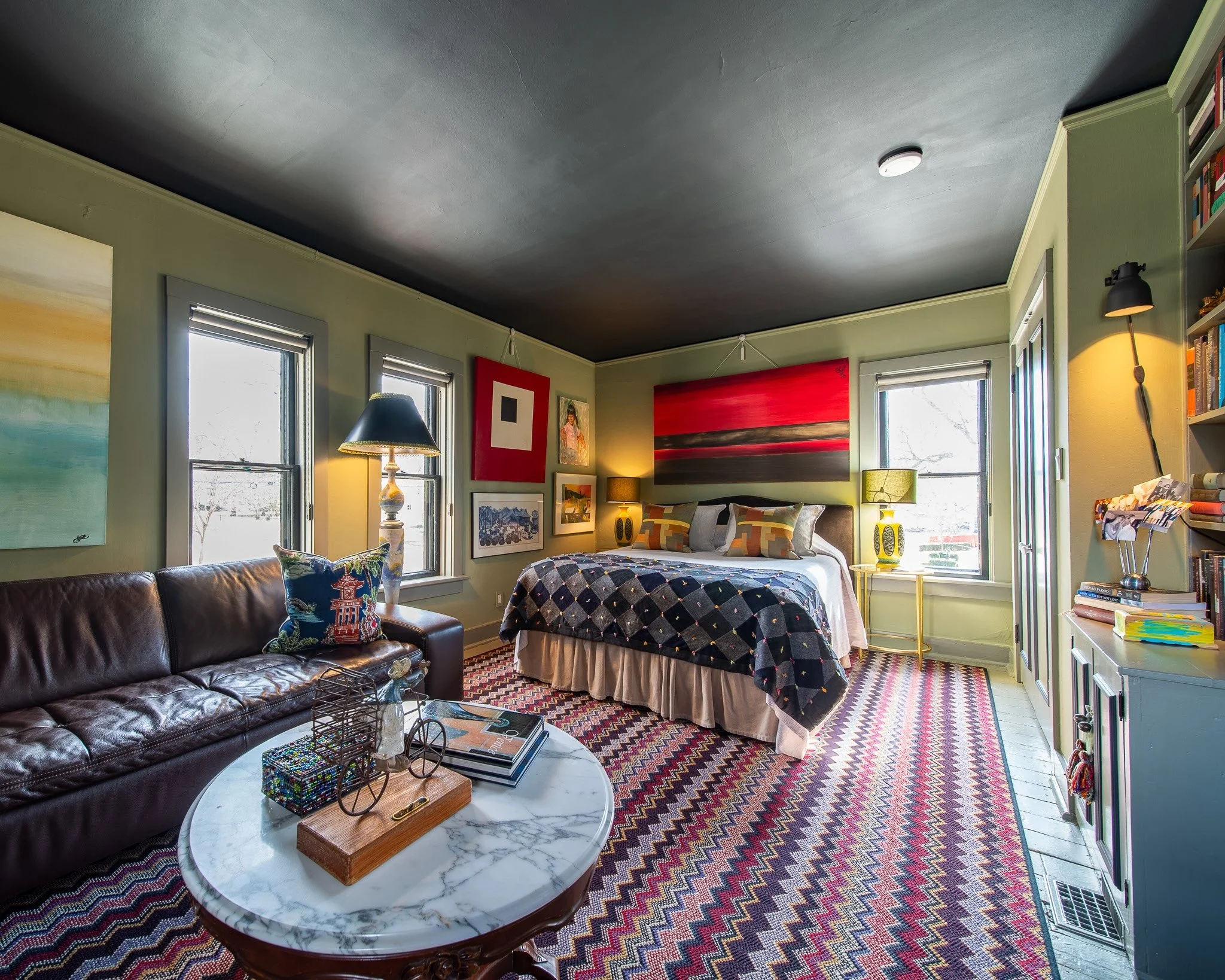 Brightly lit bedroom with green walls, large windows, colorful artwork, a bedside lamp, a plaid blanket, a decorated pillow on the bed, a red patterned rug, and a marble-top coffee table with decorative items.