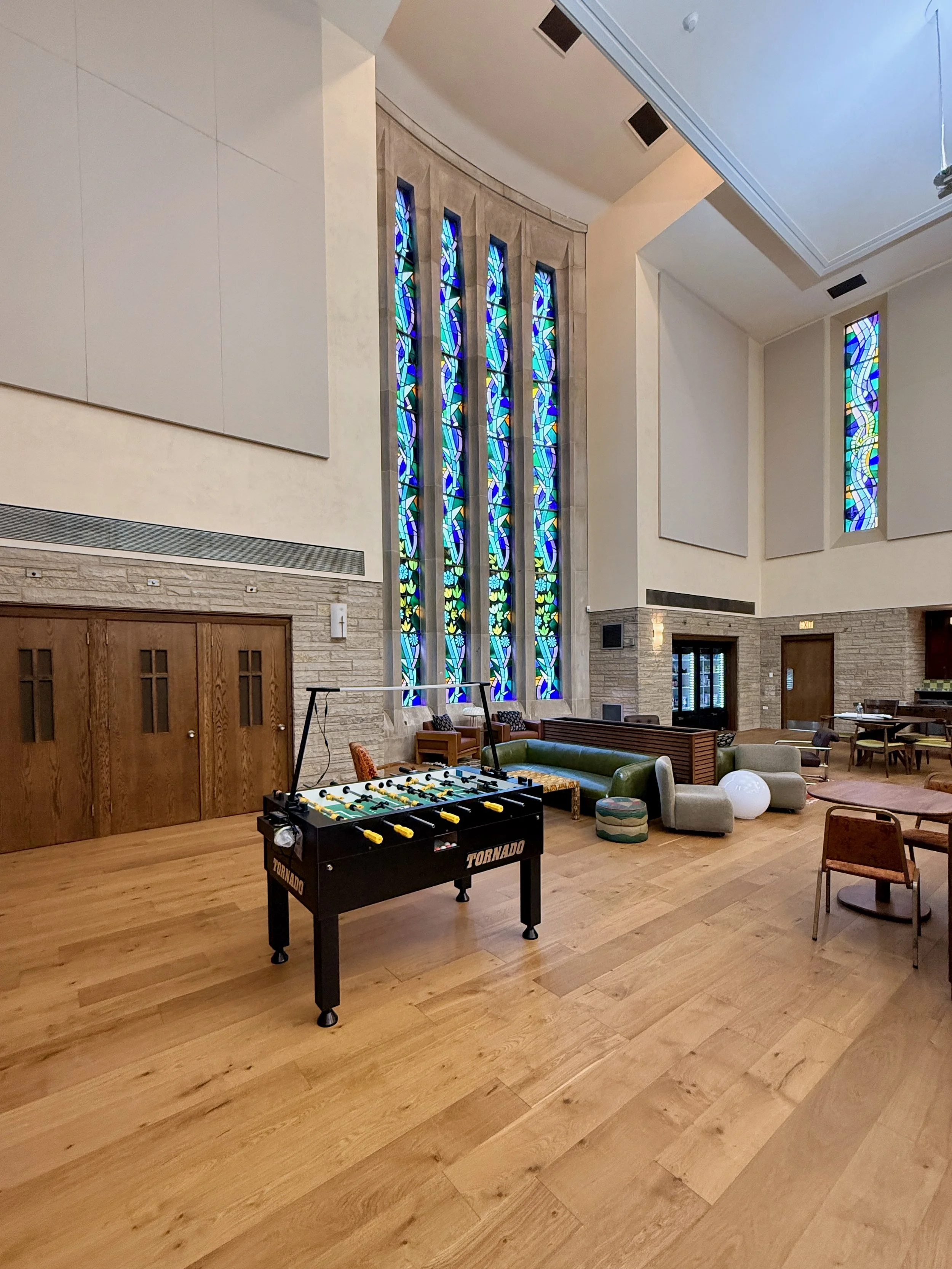 Interior view of a lounge area with large stained glass windows, wooden doors, a foosball table, and various chairs and couches.