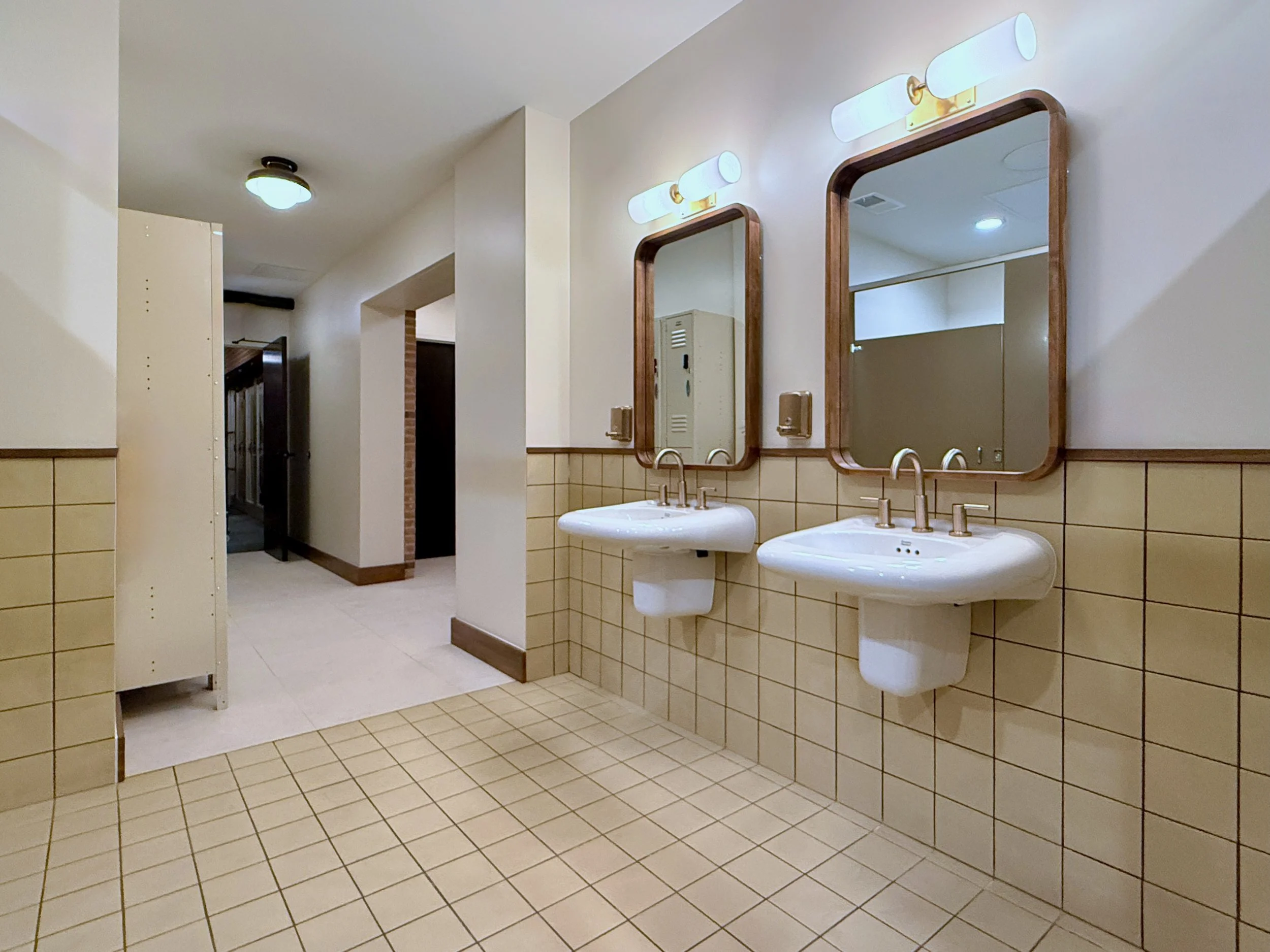 Public restroom with two sinks, two mirrors, and overhead lighting fixtures, with tiled walls and floor.