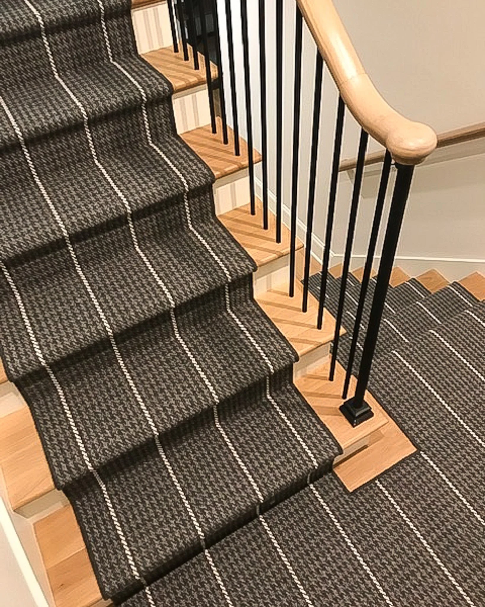 Close-up view of a staircase with black railing, beige wooden steps, and a black and beige striped carpet runner.