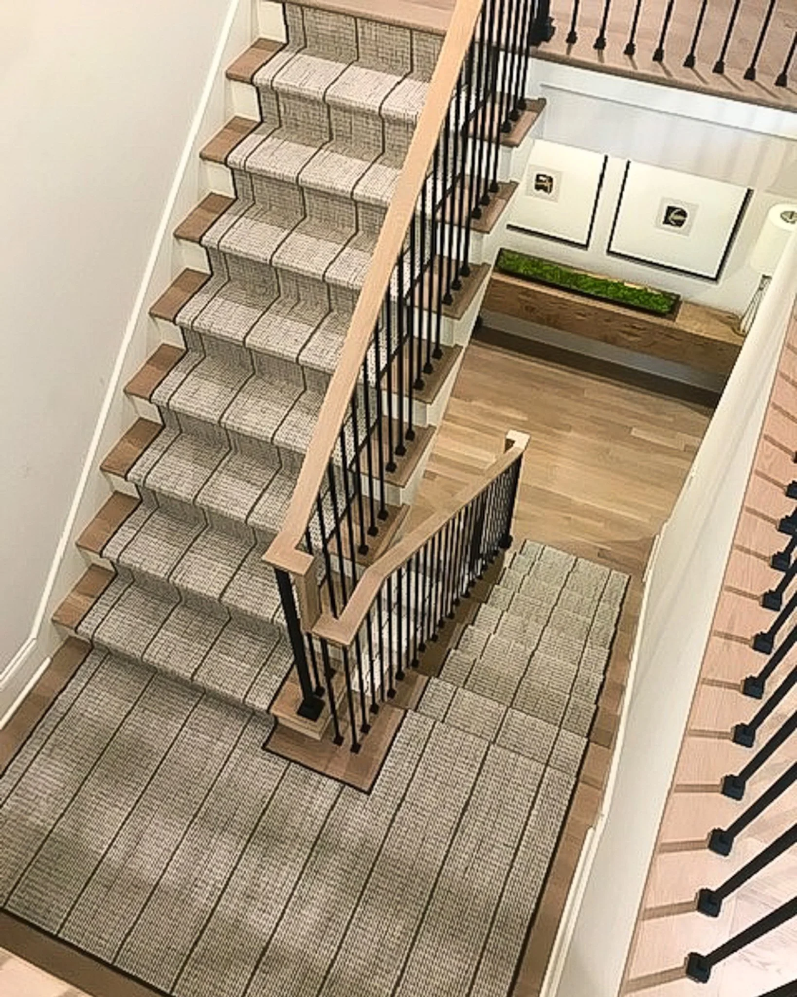 View of a staircase with carpeted steps, black metal railing, and wooden handrail, overlooking a lower level with framed pictures and a bench with a green plant.