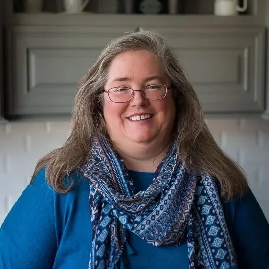 Smiling woman with glasses and long gray hair, wearing a blue shirt and patterned blue scarf, standing in front of a kitchen background.