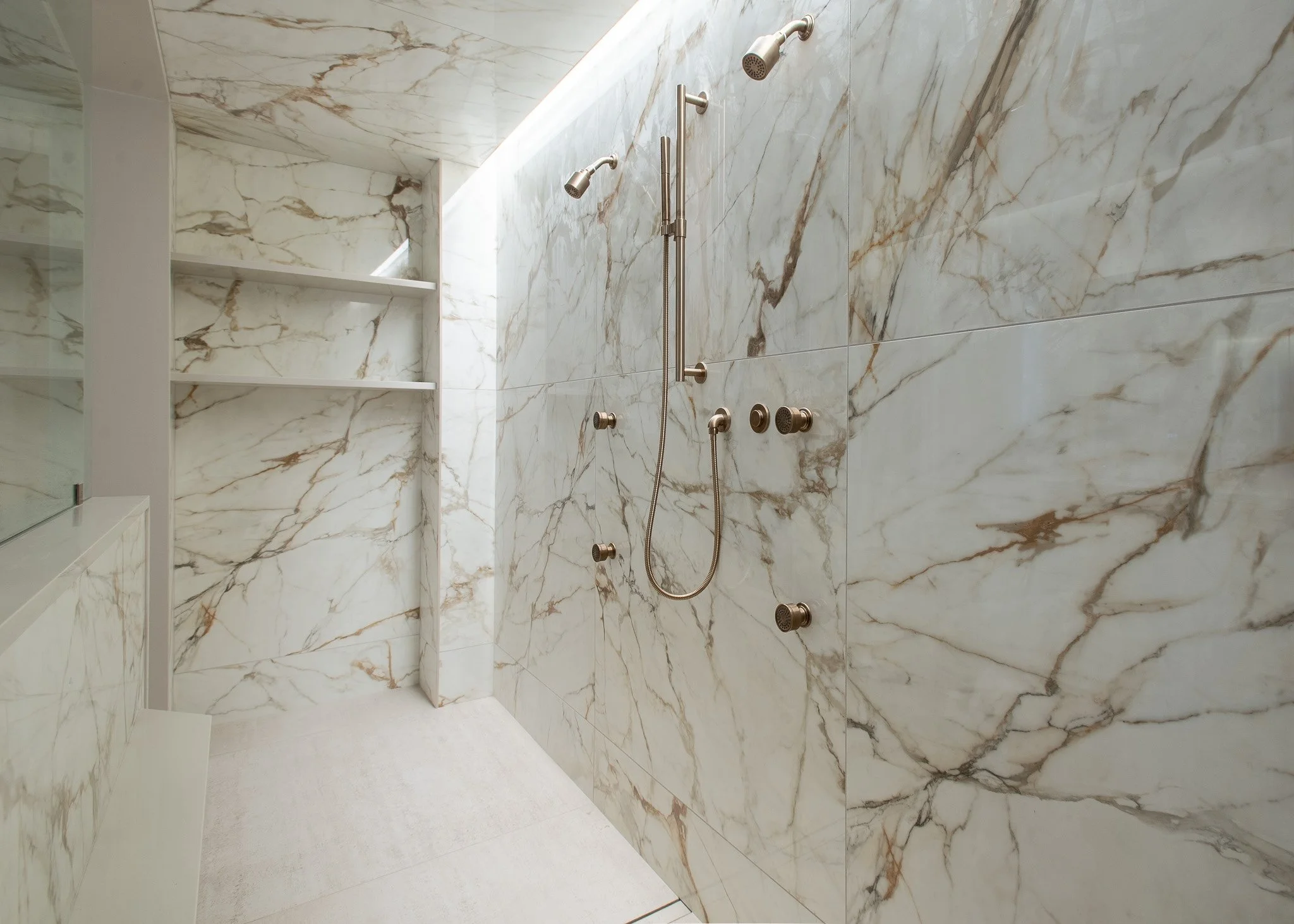 A marble shower with a built-in shelf, multiple showerheads, and a glass door.