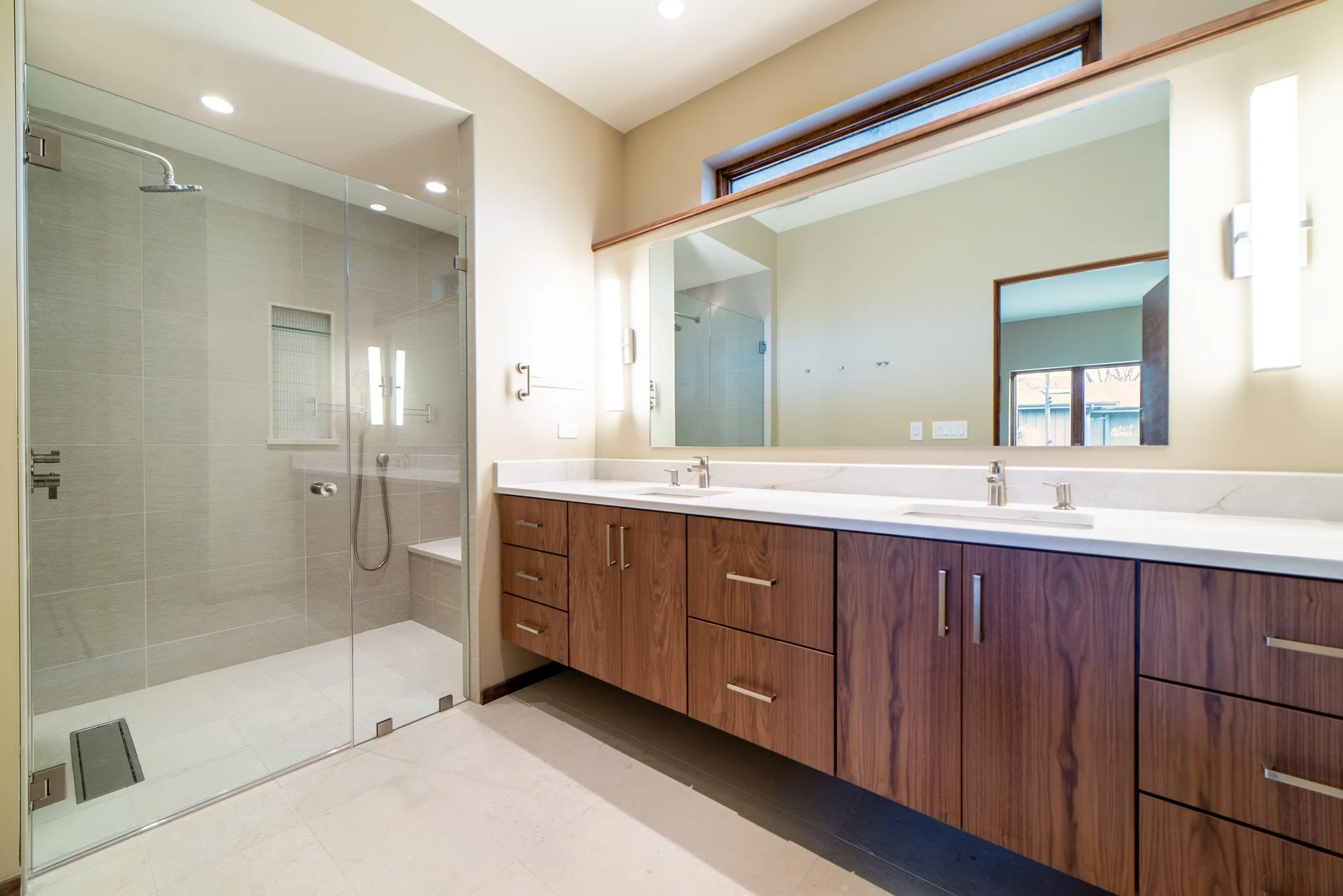 Modern bathroom with a glass-enclosed shower, wooden vanity with a double sink, large mirror, and window with wooden trim.