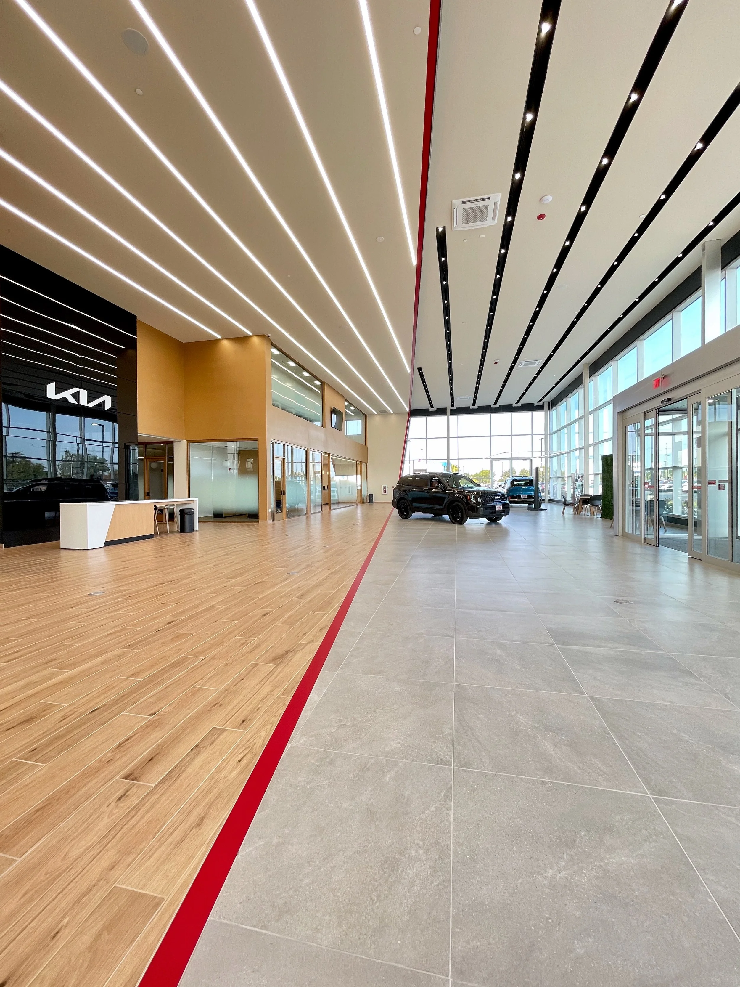 Interior of a car dealership showroom with a split design in flooring, featuring wood and tile, a black car, large windows, and modern ceiling lighting.