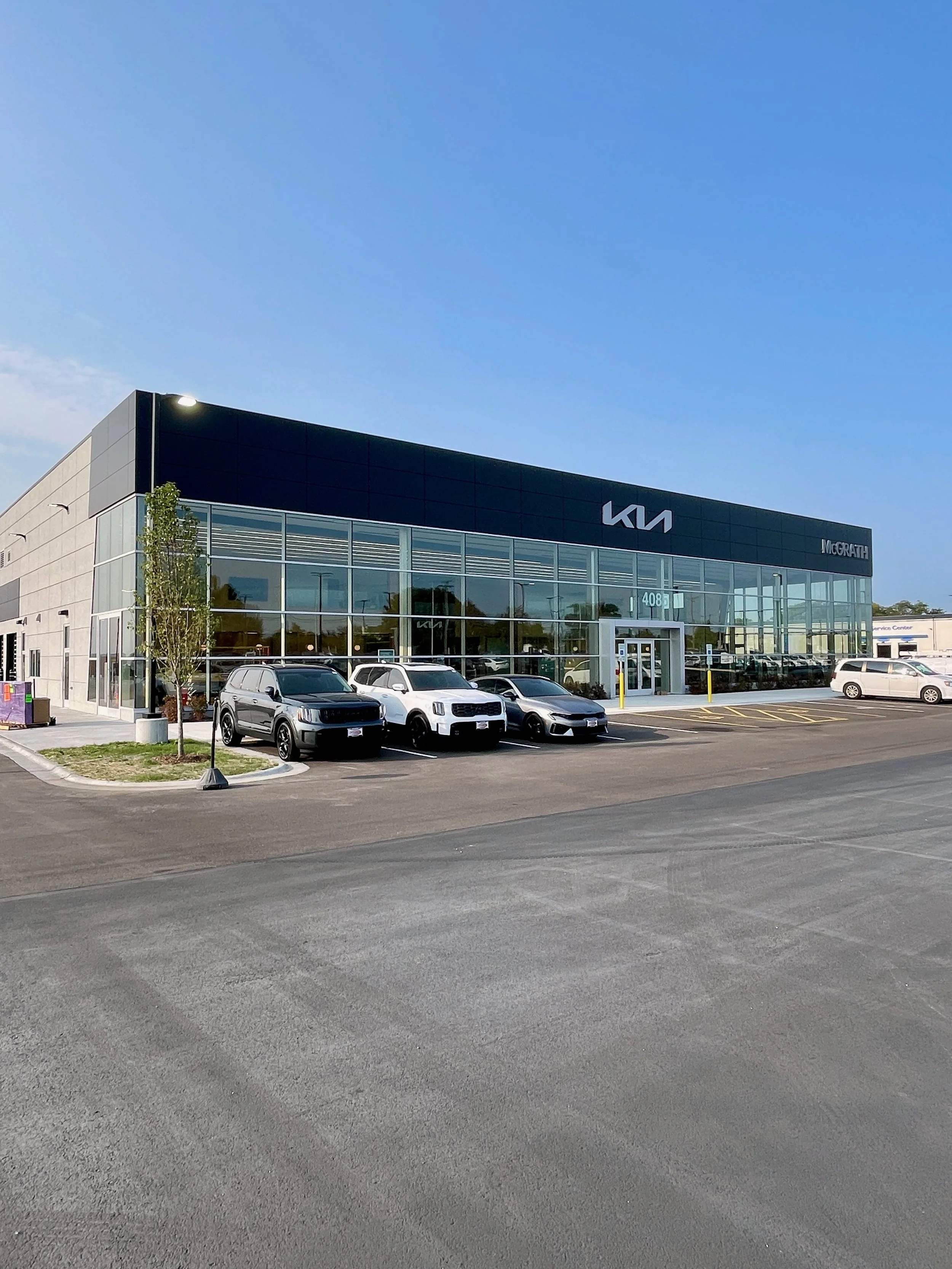 Image of a modern Kia car dealership building with a parking lot in front containing several parked vehicles, under a partly cloudy blue sky.
