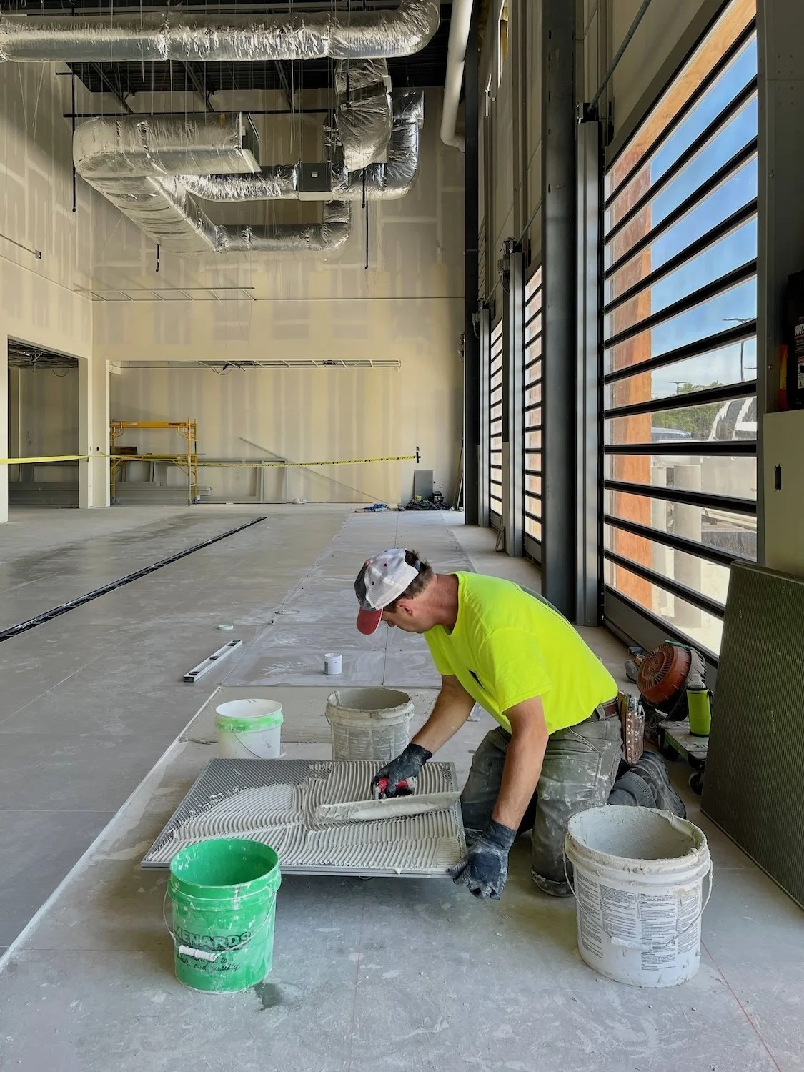 Construction worker in a bright yellow shirt and cap kneeling on the floor, applying adhesive to tiles inside a large commercial building with industrial ventilation, large windows, and construction tools and materials nearby.