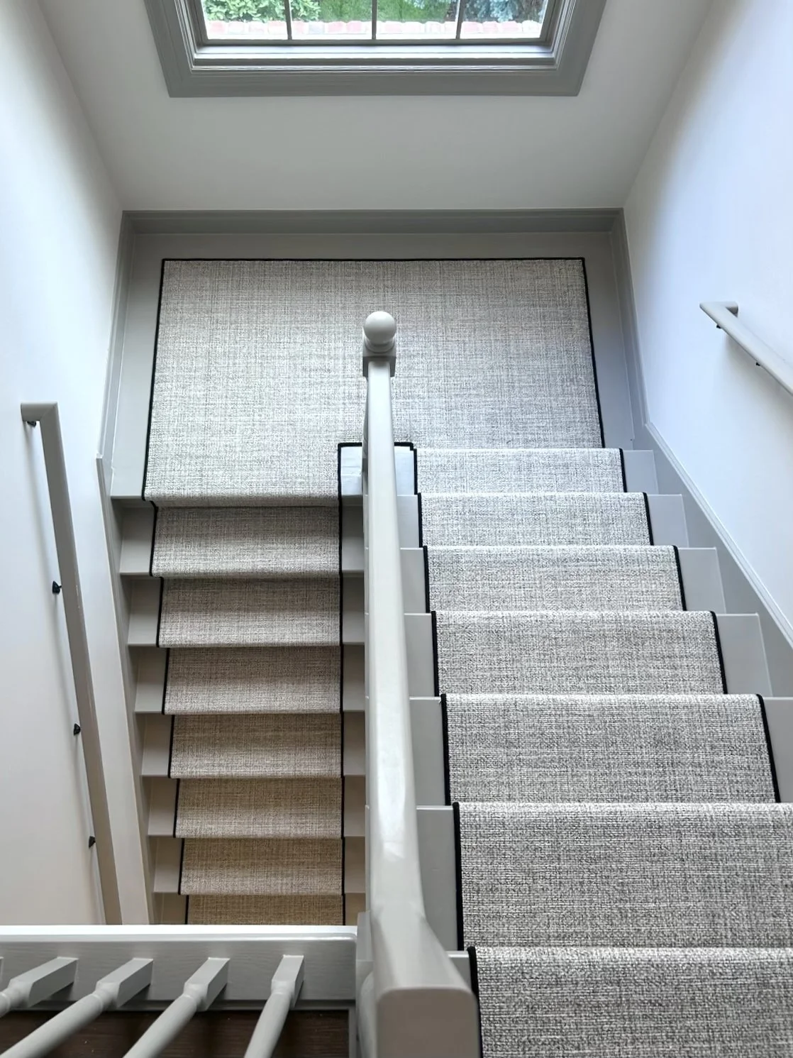 Top view of a carpeted staircase and landing seen from above, with a window at the top and white walls on either side.