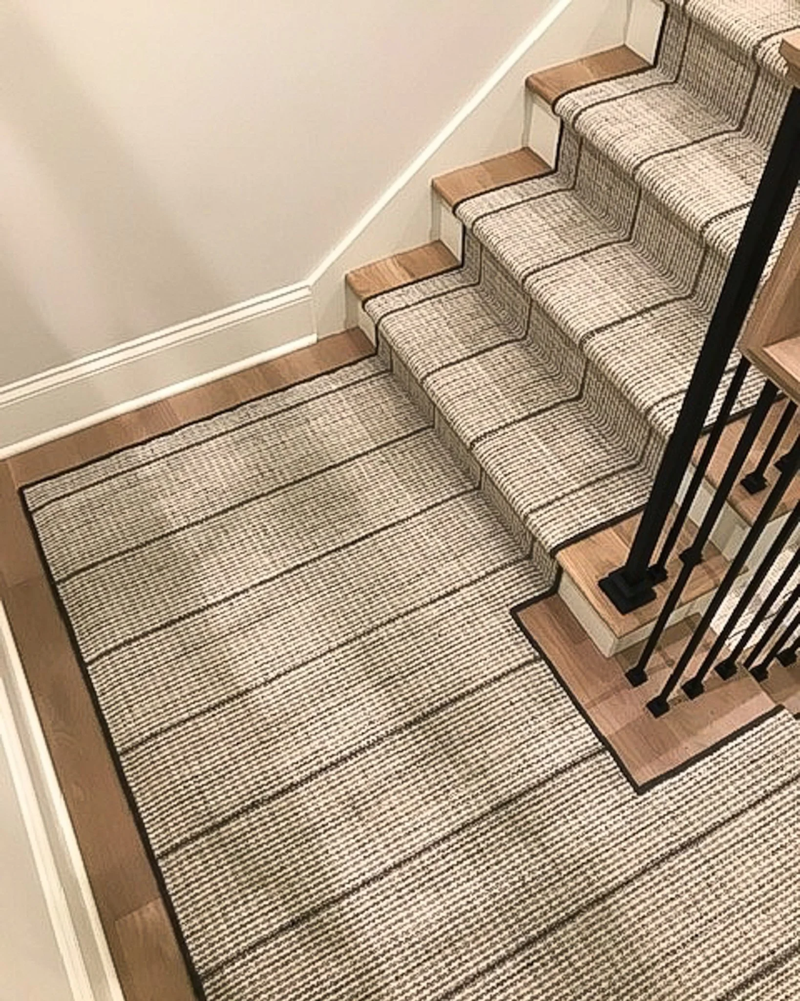 A staircase with a patterned carpet runner, black iron railing, and wooden steps, next to beige walls and baseboards.