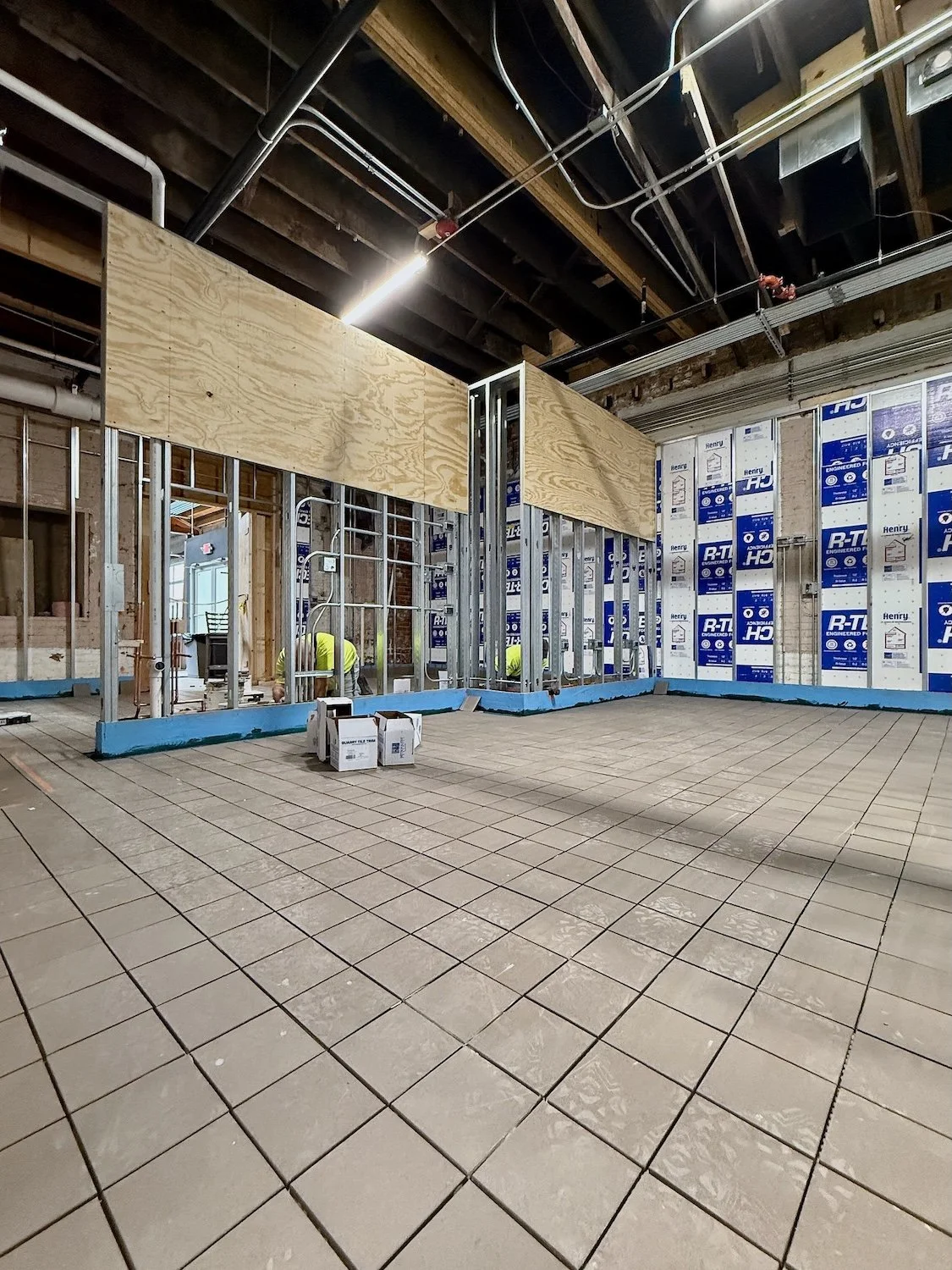 Indoor construction site with workers installing framing for walls, featuring tiled floor and exposed ceiling with visible pipes and wiring.