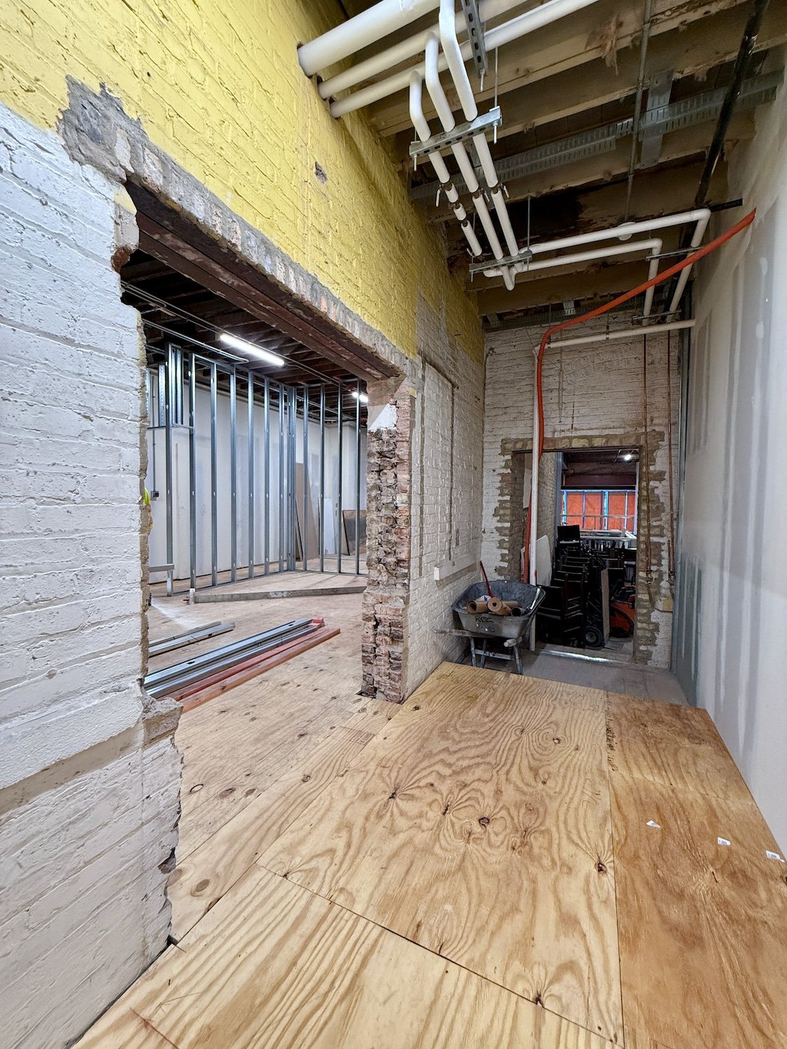 Interior of a building under construction with exposed brick walls, visible plumbing pipes, and framing for new walls. There is a wheelbarrow with construction materials and wood flooring partially installed.