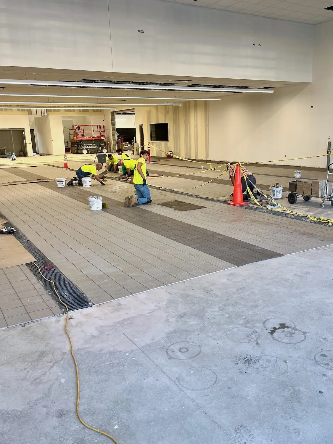 Workers installing or repairing floor tiles in a large indoor space, with construction cones, caution tape, and construction tools present.