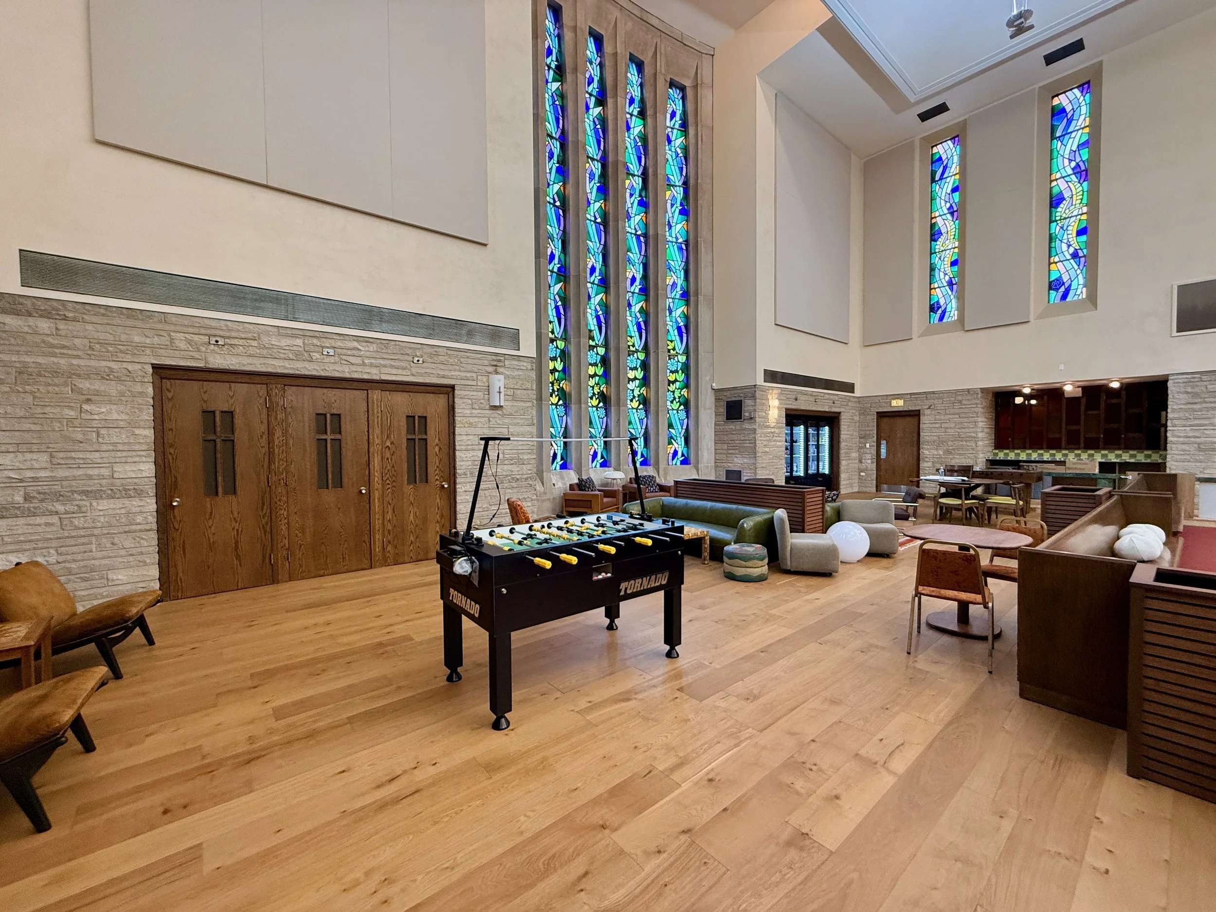 A spacious, well-lit room with large stained glass windows, wooden flooring, comfortable seating, a foosball table, and various furniture pieces, suggesting a lounge or recreational area.