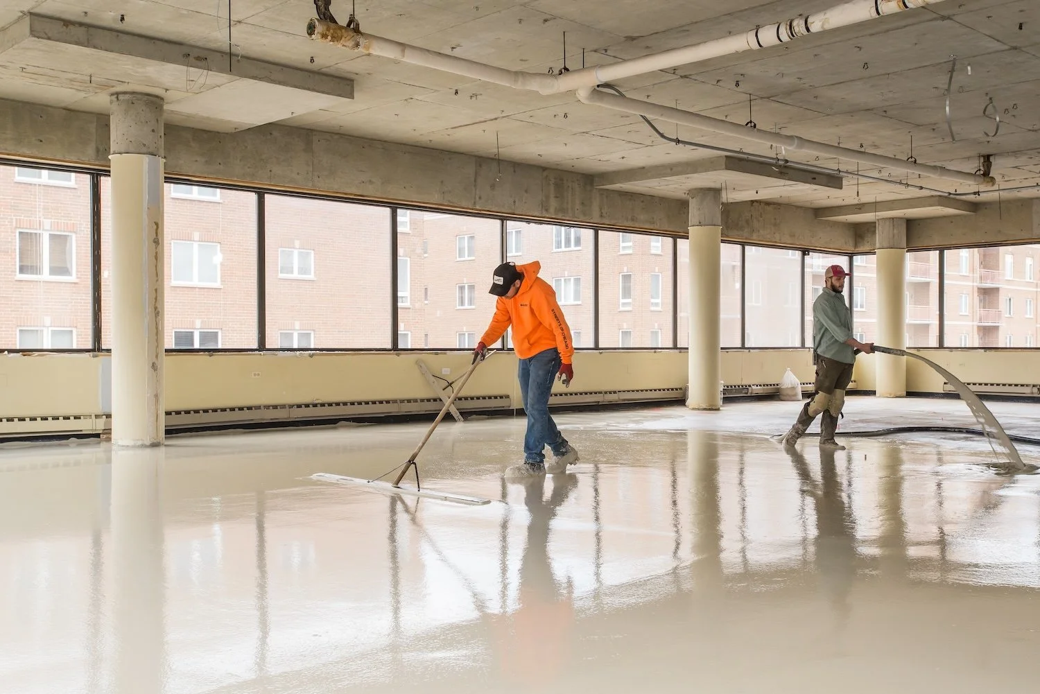 Two construction workers are pouring and leveling concrete on a large indoor floor with large windows in the background.
