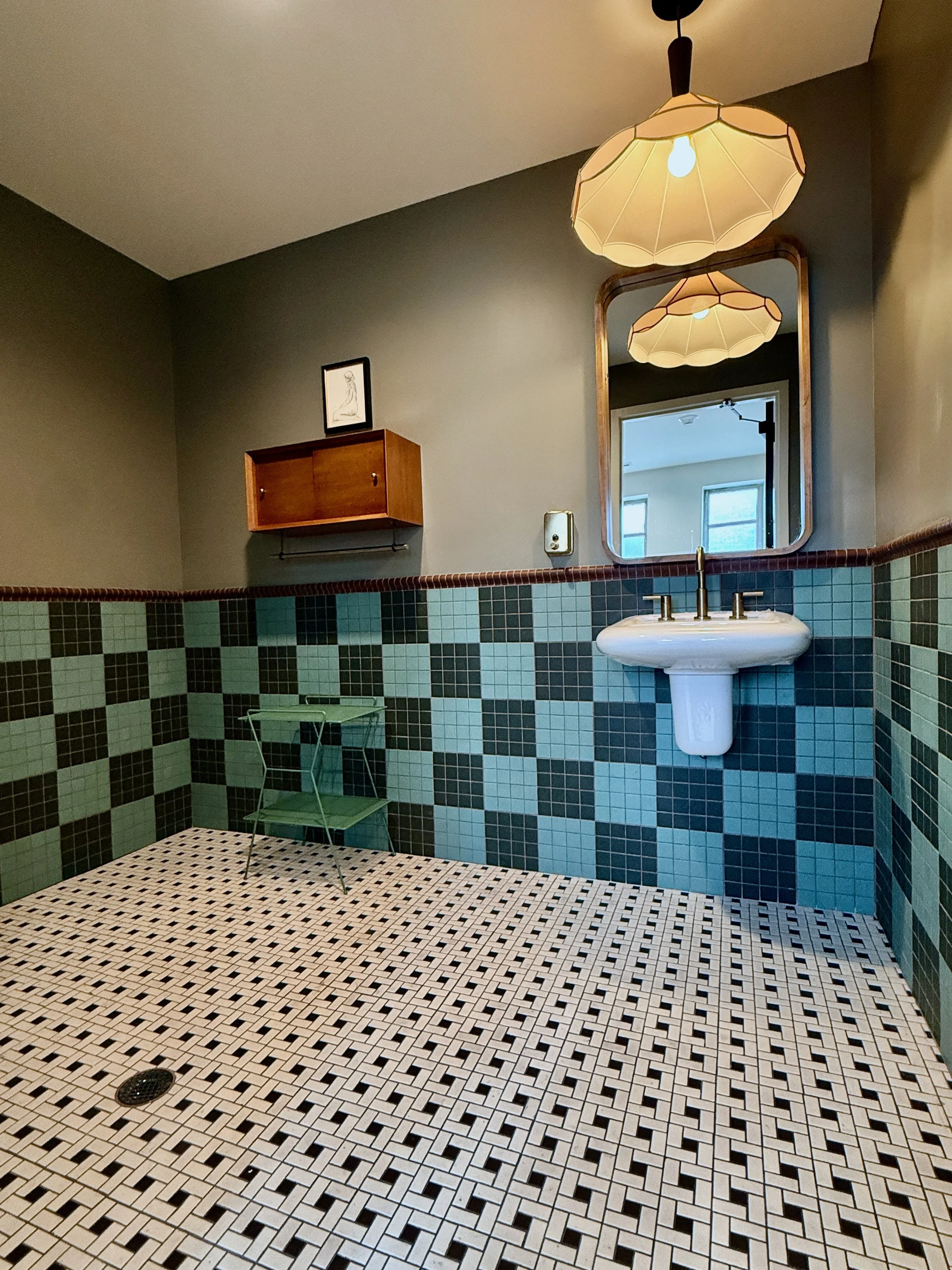 A bathroom with white and black square tile floor, green and black tiled lower wall, dark green upper wall, wooden wall cabinet, large mirror, hanging ceiling light, white pedestal sink, and small green wire shelf at the corner.