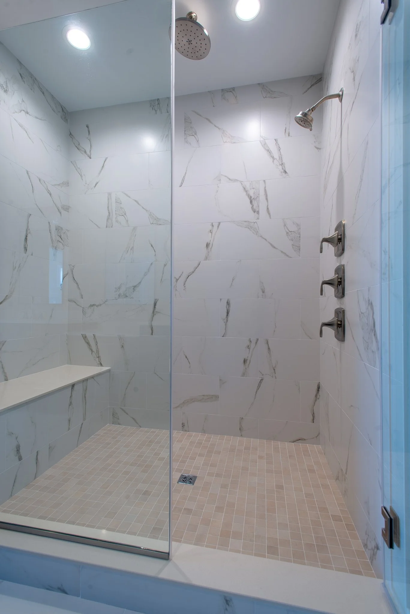 A walk-in shower with marble walls, a built-in bench, a rainfall showerhead on the ceiling, and a handheld showerhead with three controls.
