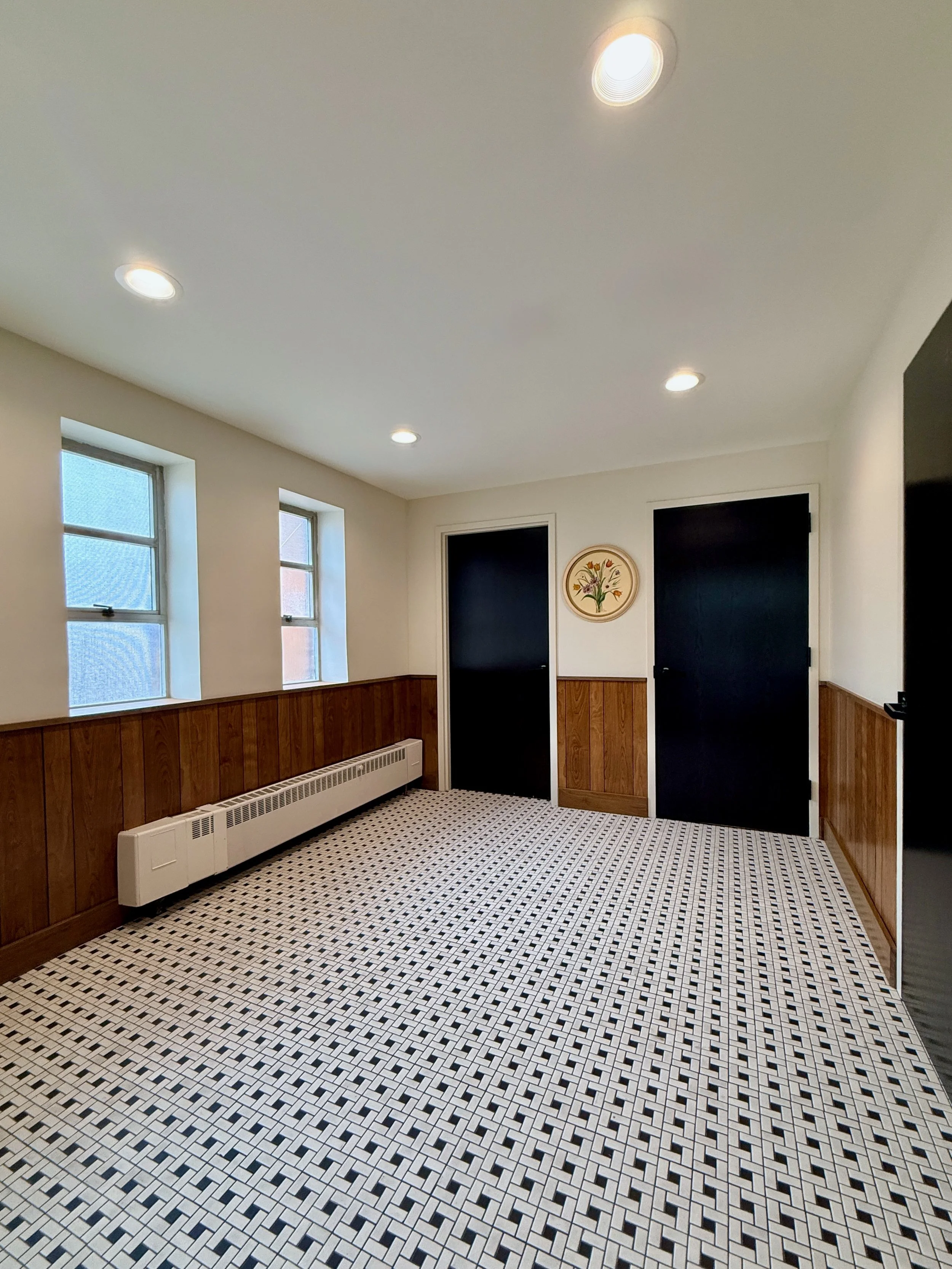 A room with three windows, a white ceiling with recessed lights, and a black door. The lower half of the walls is covered with wooden paneling, and the floor has a black and white geometric tile pattern.