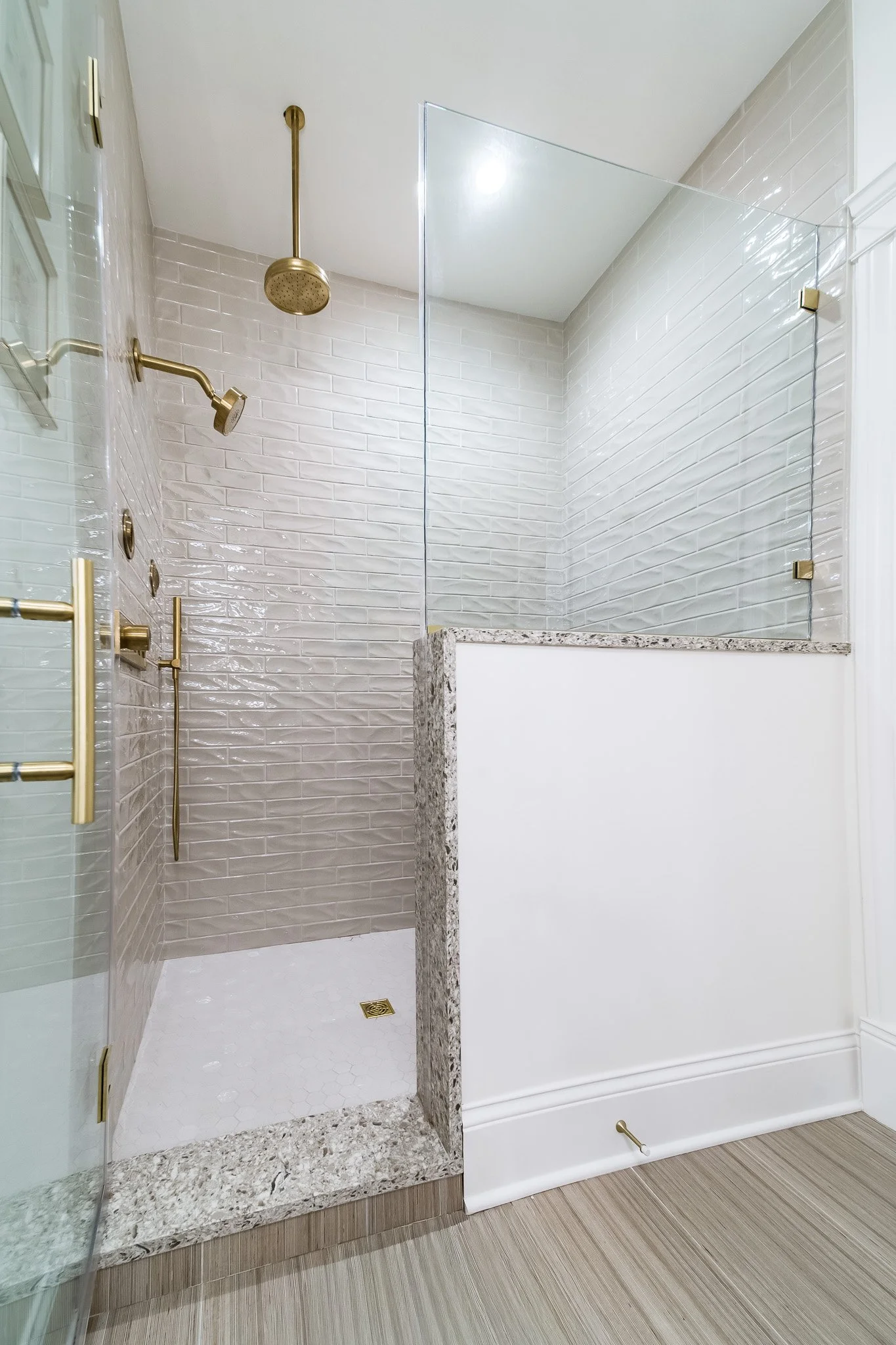 Modern shower area with gold fixtures, a glass partition, white subway tile walls, a marble threshold, and a white tiled floor.