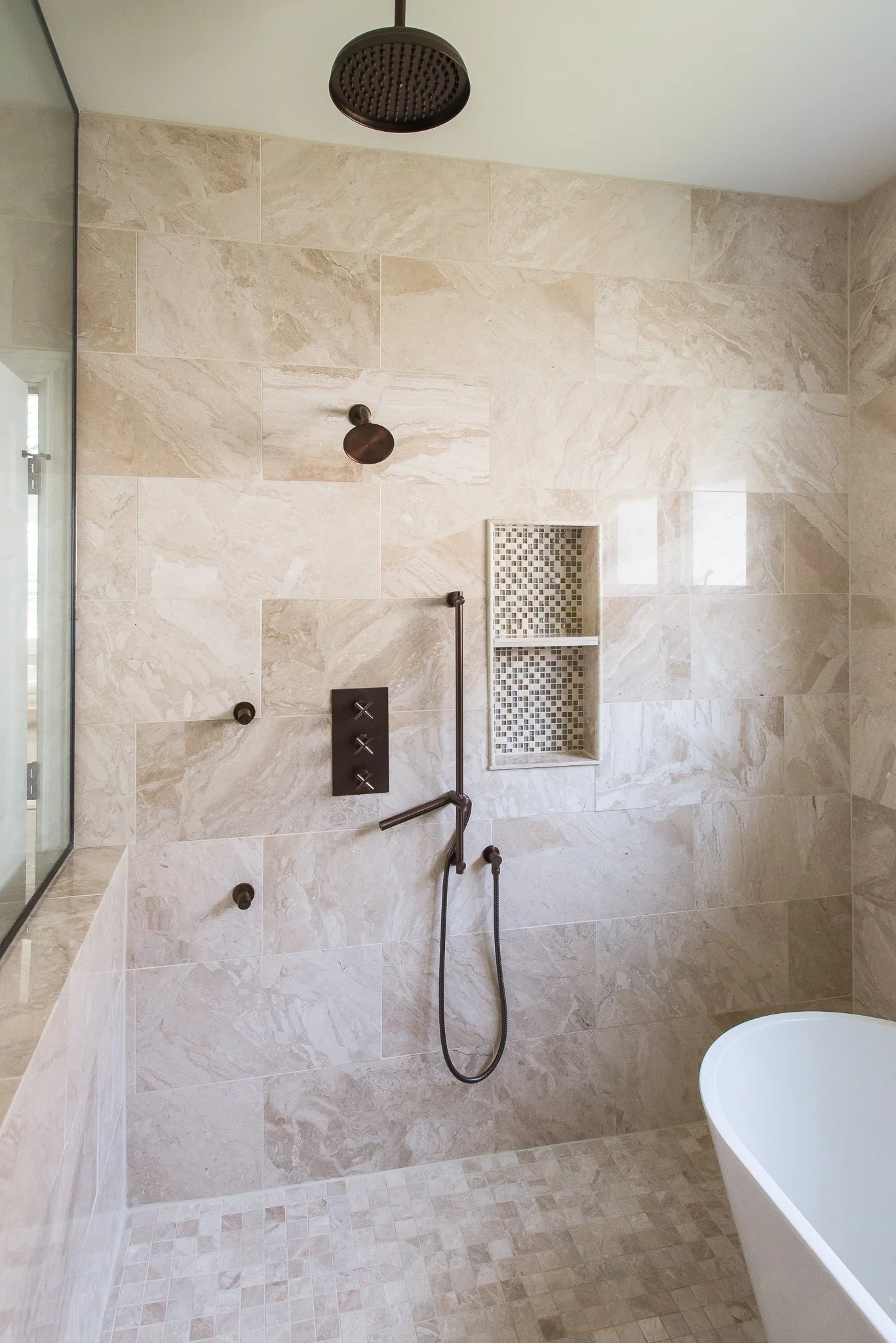 A modern shower with beige marble tiles, a rain showerhead, a handheld shower, a small built-in niche with mosaic tiles, and a partial view of a freestanding bathtub.