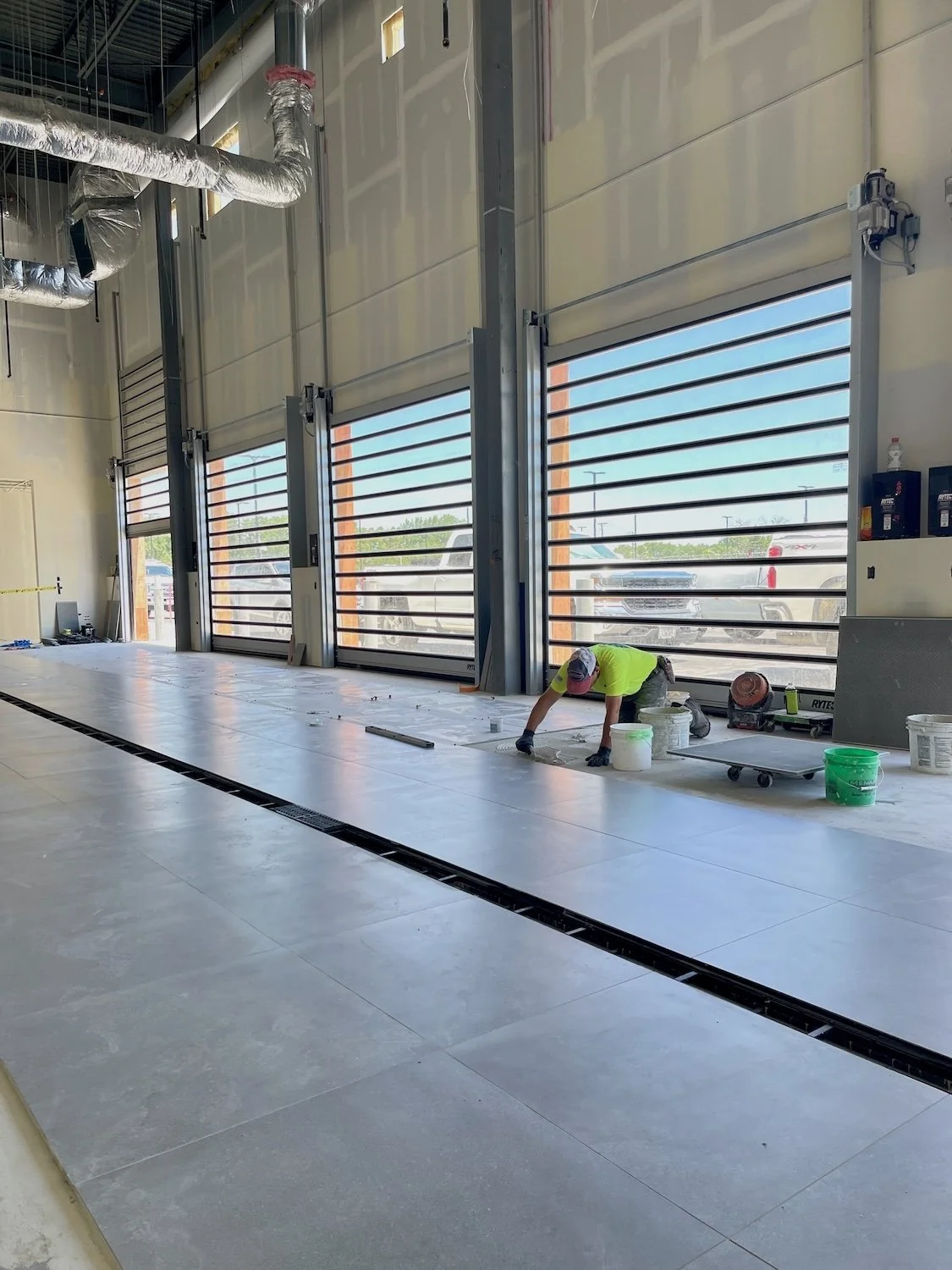 Worker kneeling on the floor installing large tiles inside a spacious commercial building with large garage-style windows and visible HVAC ductwork.