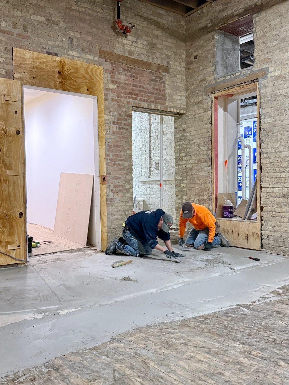 Two construction workers kneel on the floor of an unfinished room with exposed brick walls, working on pouring or smoothing concrete. The room has open doorways and a window opening, with construction materials and tools around them.