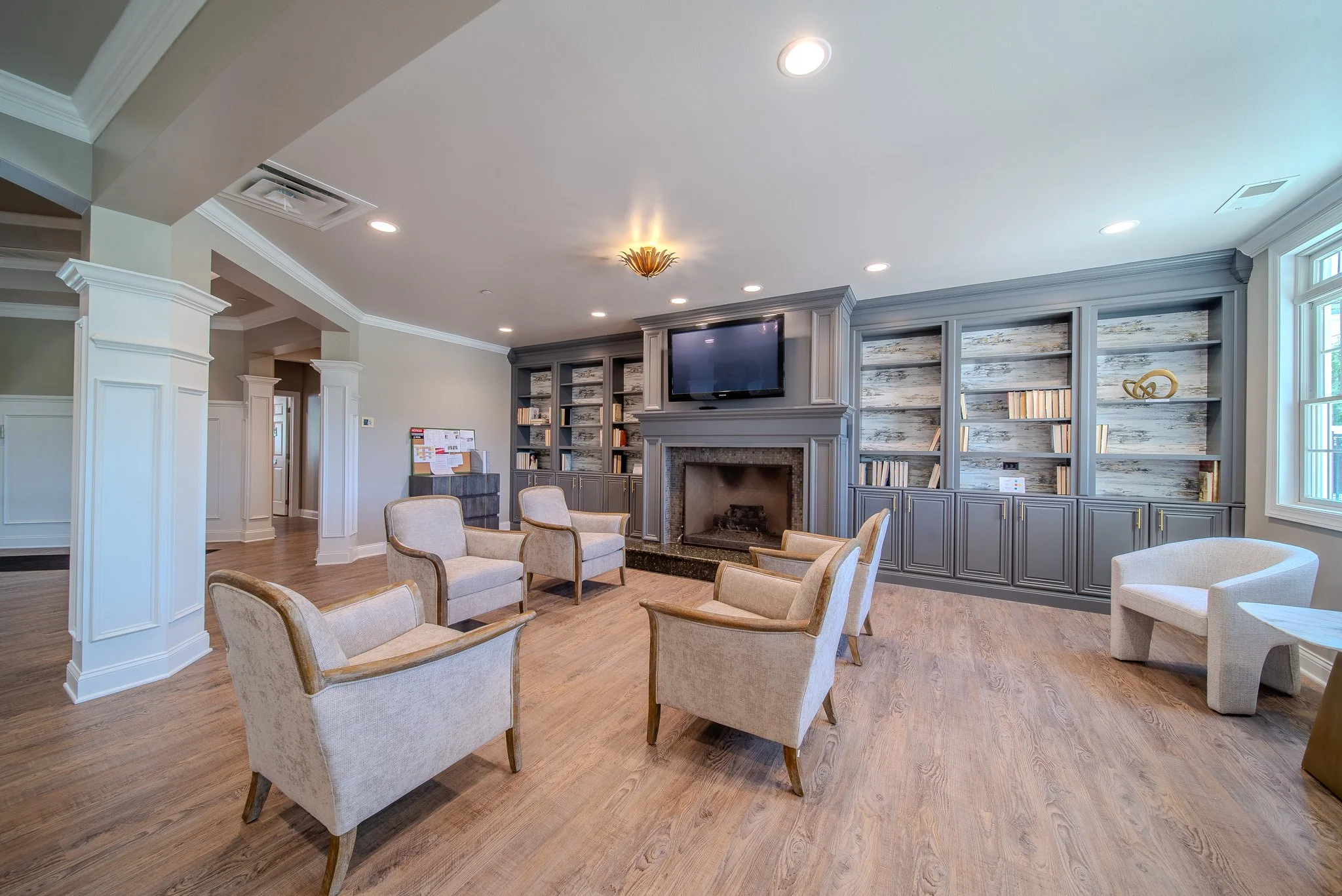 A spacious living room with multiple beige armchairs arranged in a circle around a fireplace and a mounted flat-screen TV above it. The room features built-in gray bookshelves with books and decorative items, and has large windows allowing natural li
