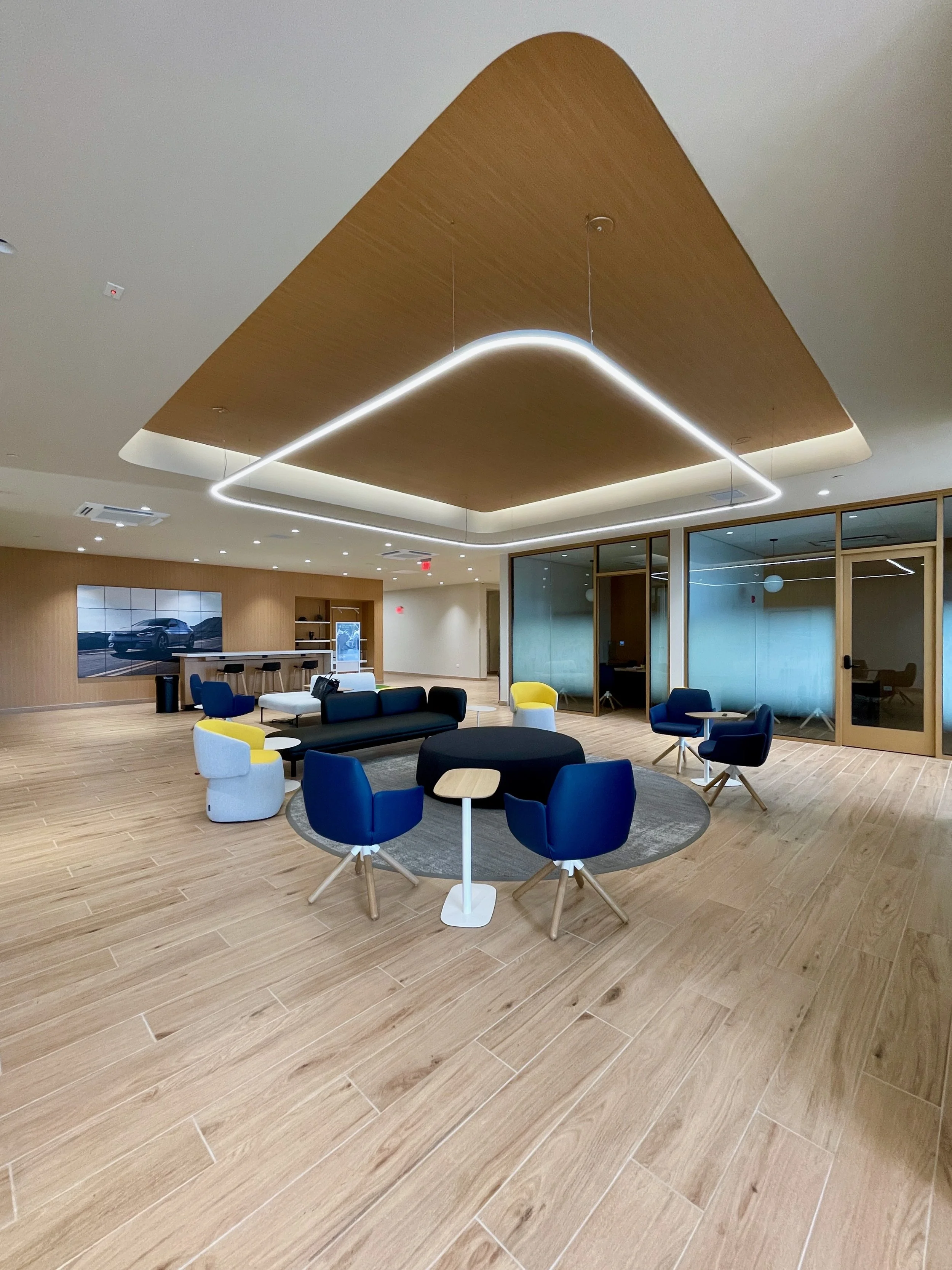 Modern office lounge with a wooden ceiling feature, a circular black rug, and various blue, yellow, and white chairs and sofas, with glass partitions and a TV screen showing a car in the background.